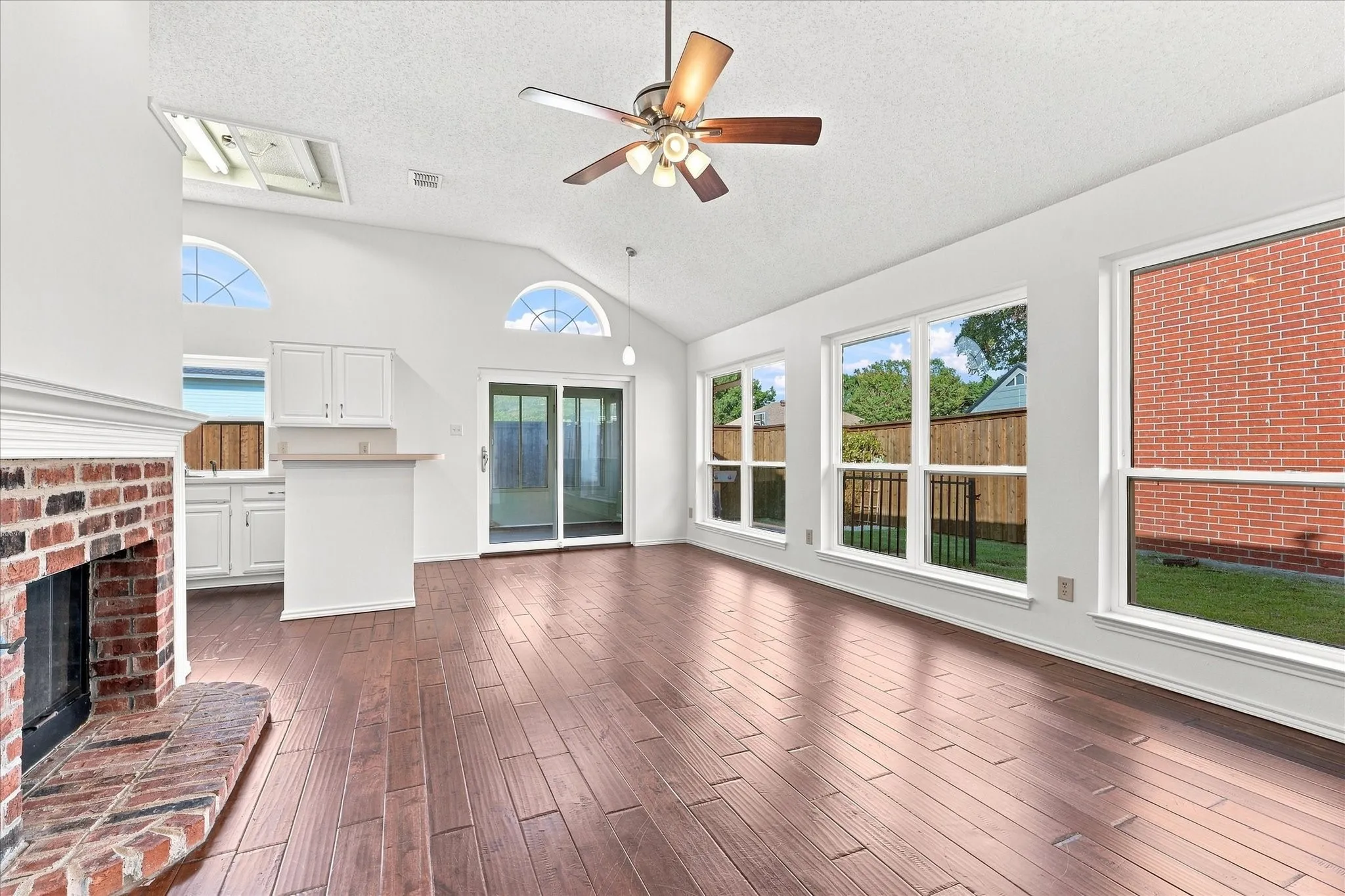 Vaulted ceiling with a ceiling fan, brick fireplace, wood-finish flooring, white cabinetry, and sliding glass doors