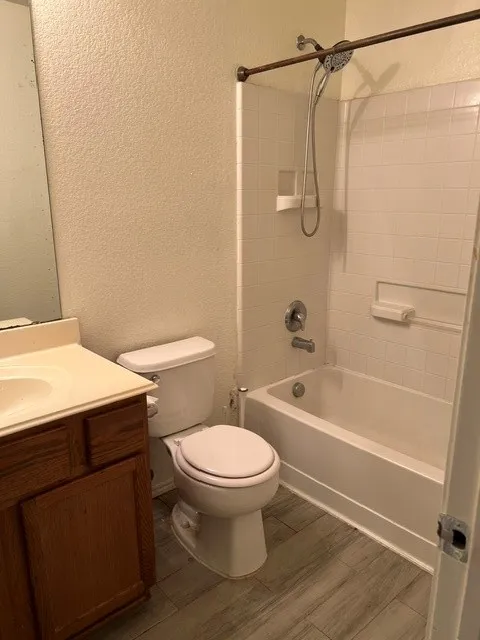 Wood-finish flooring, white tiled shower surround, white bathtub, vanity with integrated sink, and a toilet