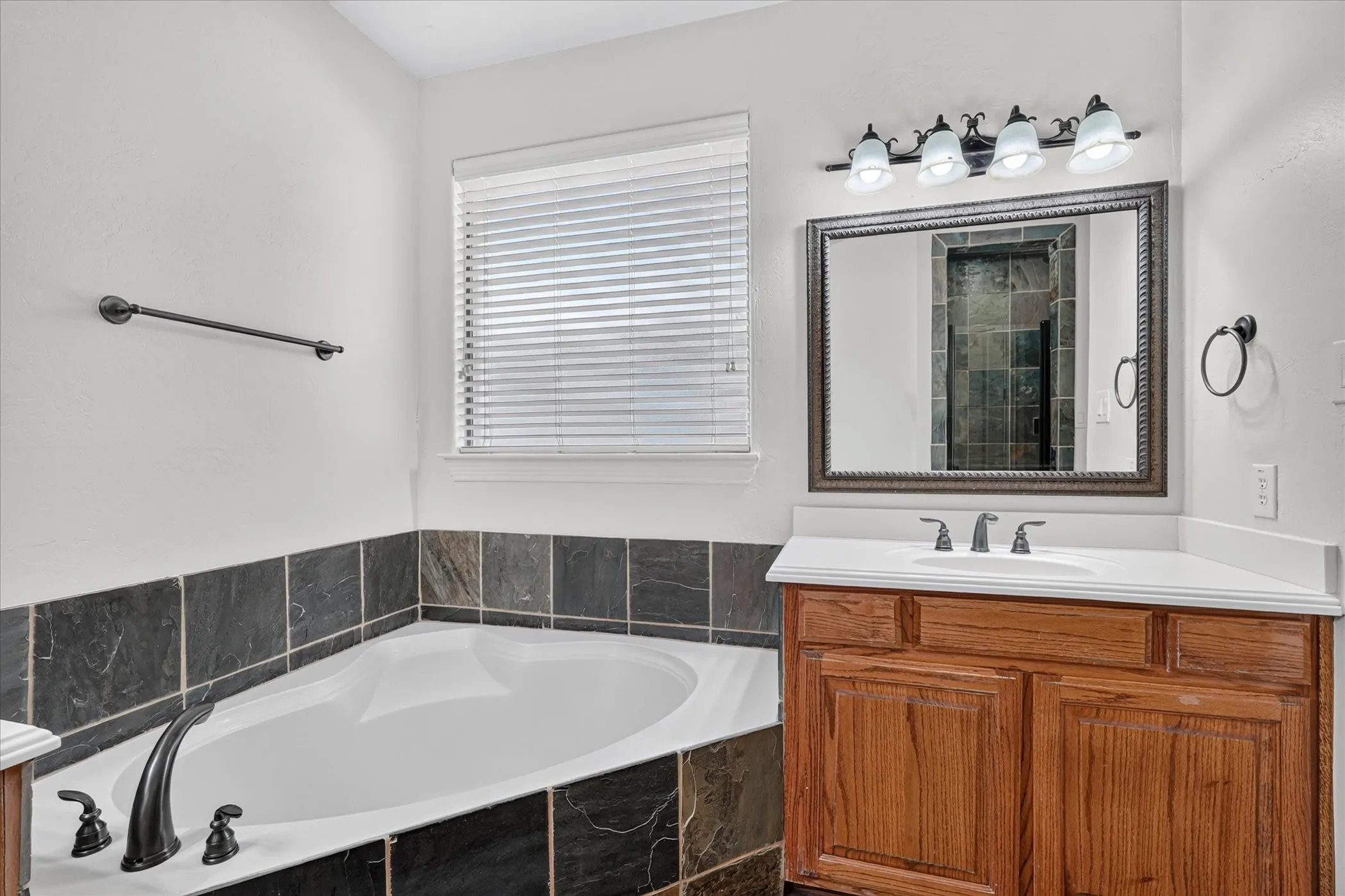 Built-in soaking tub with dark tile surround, wood-finish vanity with white countertop, framed mirror, and four-light vanity fixture