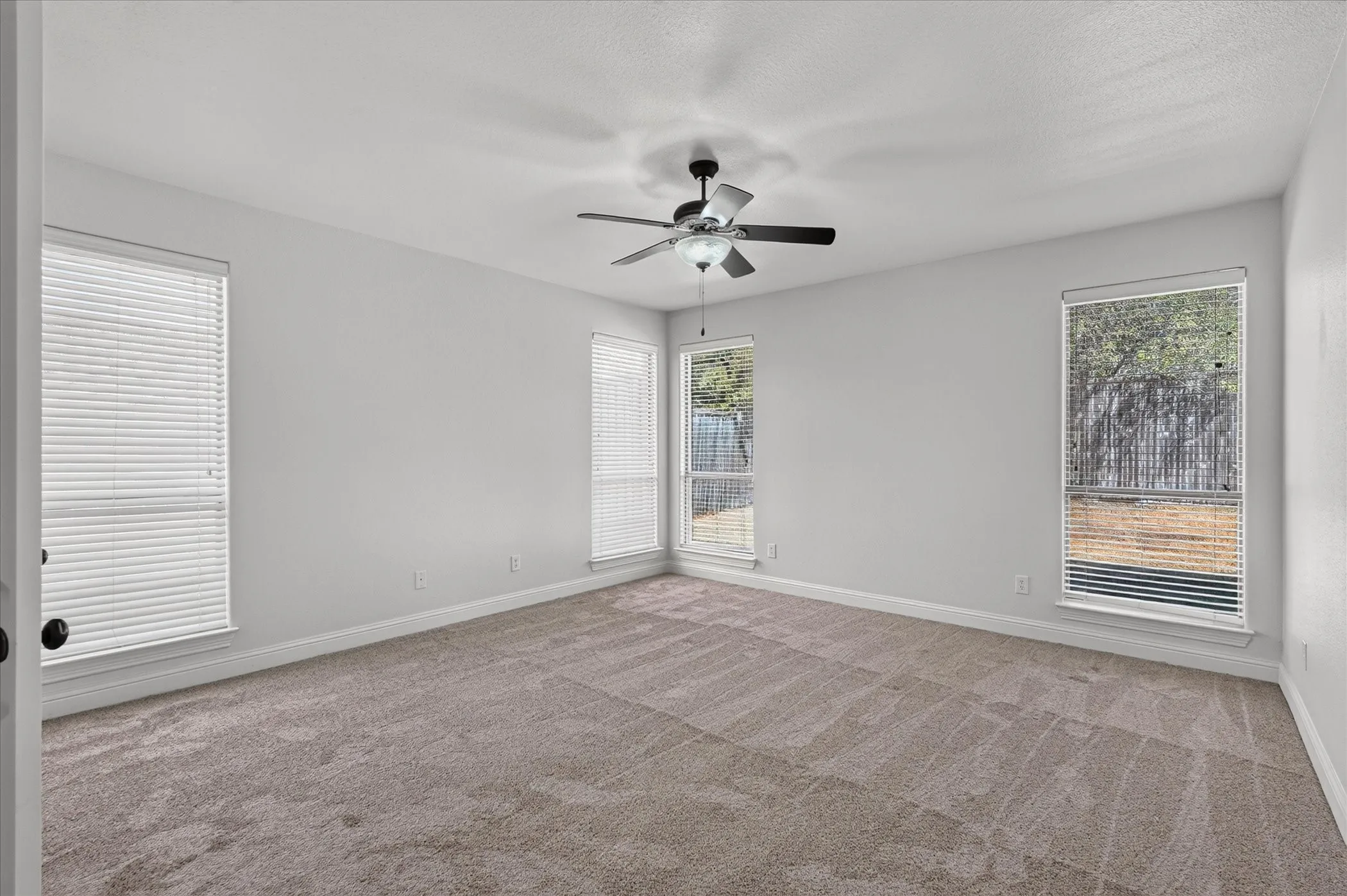 Spacious room featuring neutral carpet flooring, three windows with blinds, and a ceiling fan with integrated lighting