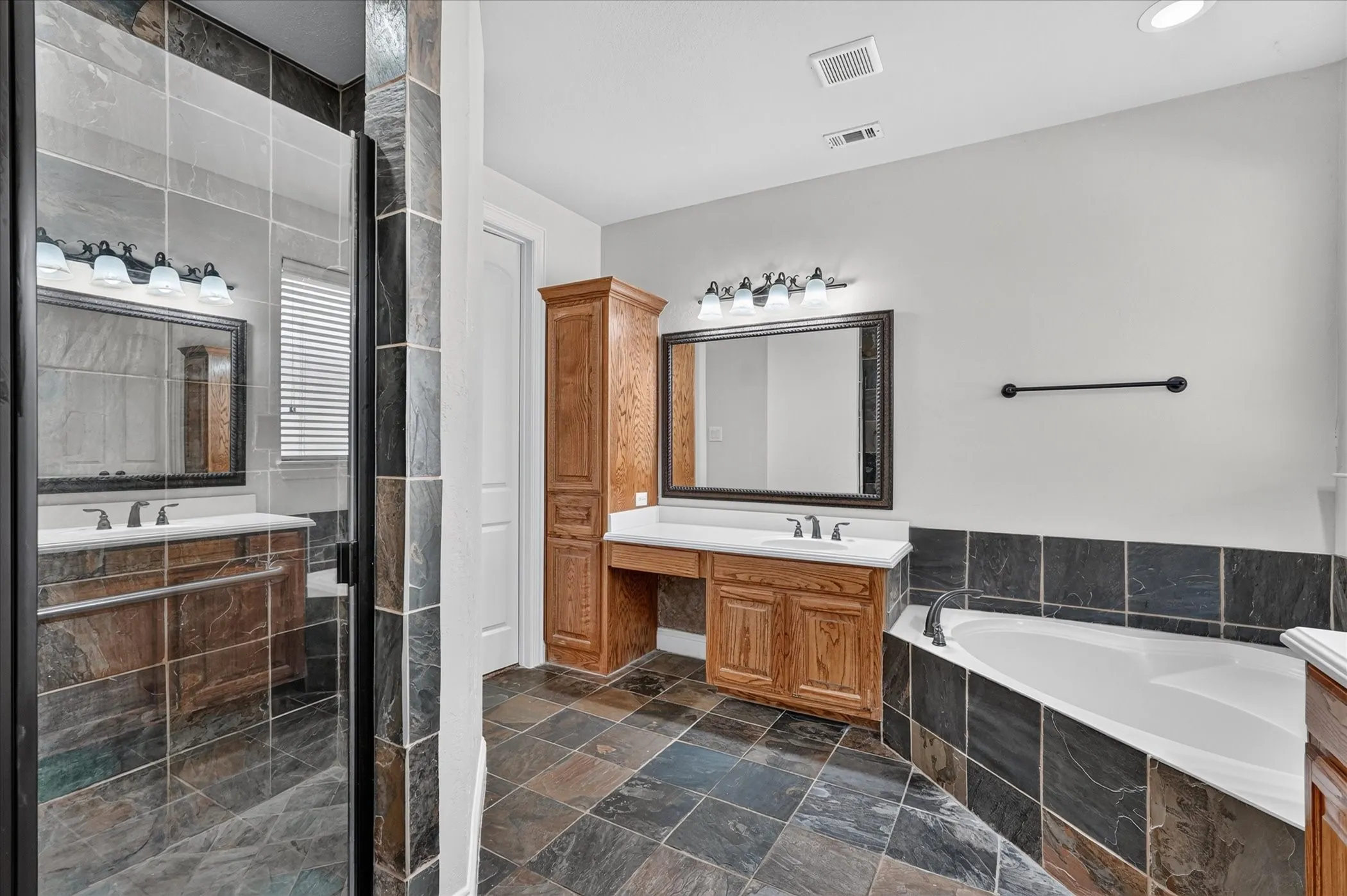 Spacious bathroom with stone-finish tile flooring, a built-in soaking tub, wood cabinetry with white countertops, a large framed mirror, and a glass-enclosed shower