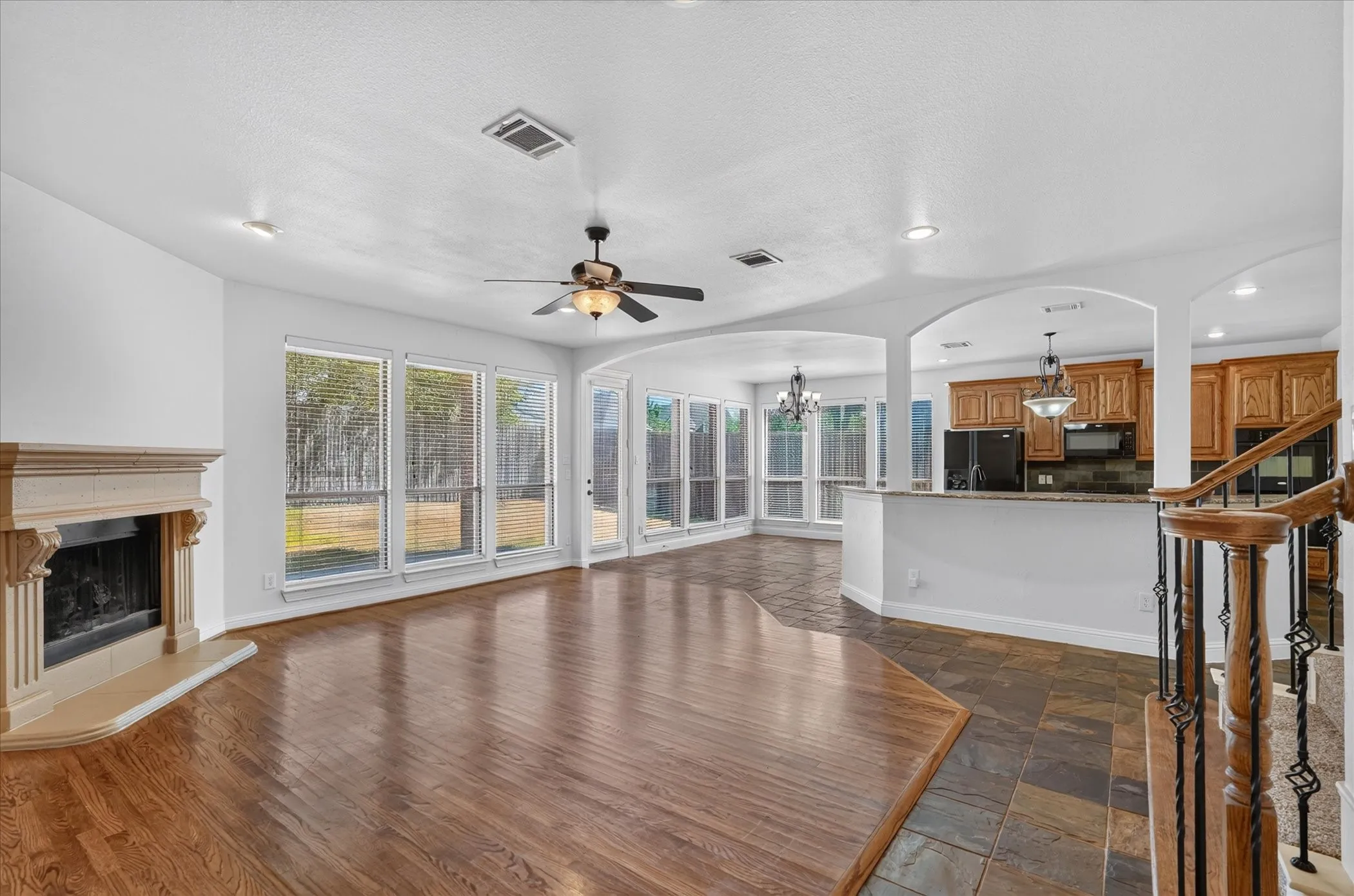 Hardwood flooring, fireplace with ornate mantel, multiple large windows, ceiling fan, and staircase with wrought iron balusters