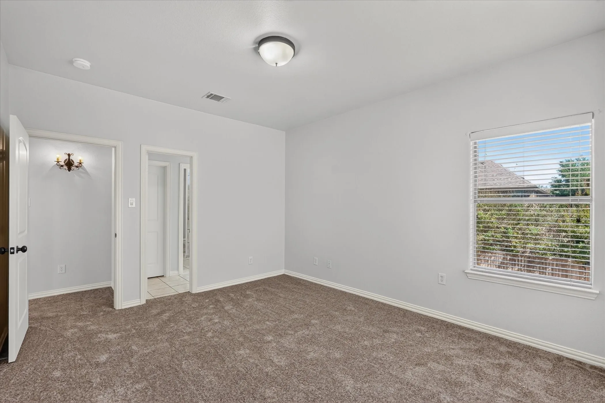 Neutral wall paint, brown carpet flooring, flush mount ceiling light, window with horizontal blinds, and a built-in wall sconce