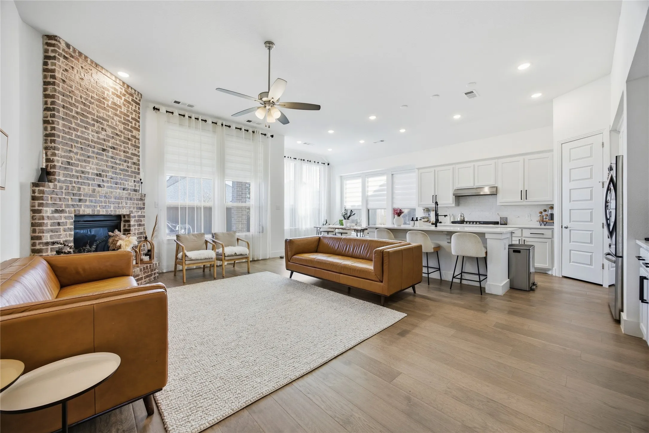 Spacious open-concept living area featuring a brick fireplace, wood-finish flooring, a ceiling fan, recessed lighting, and large windows