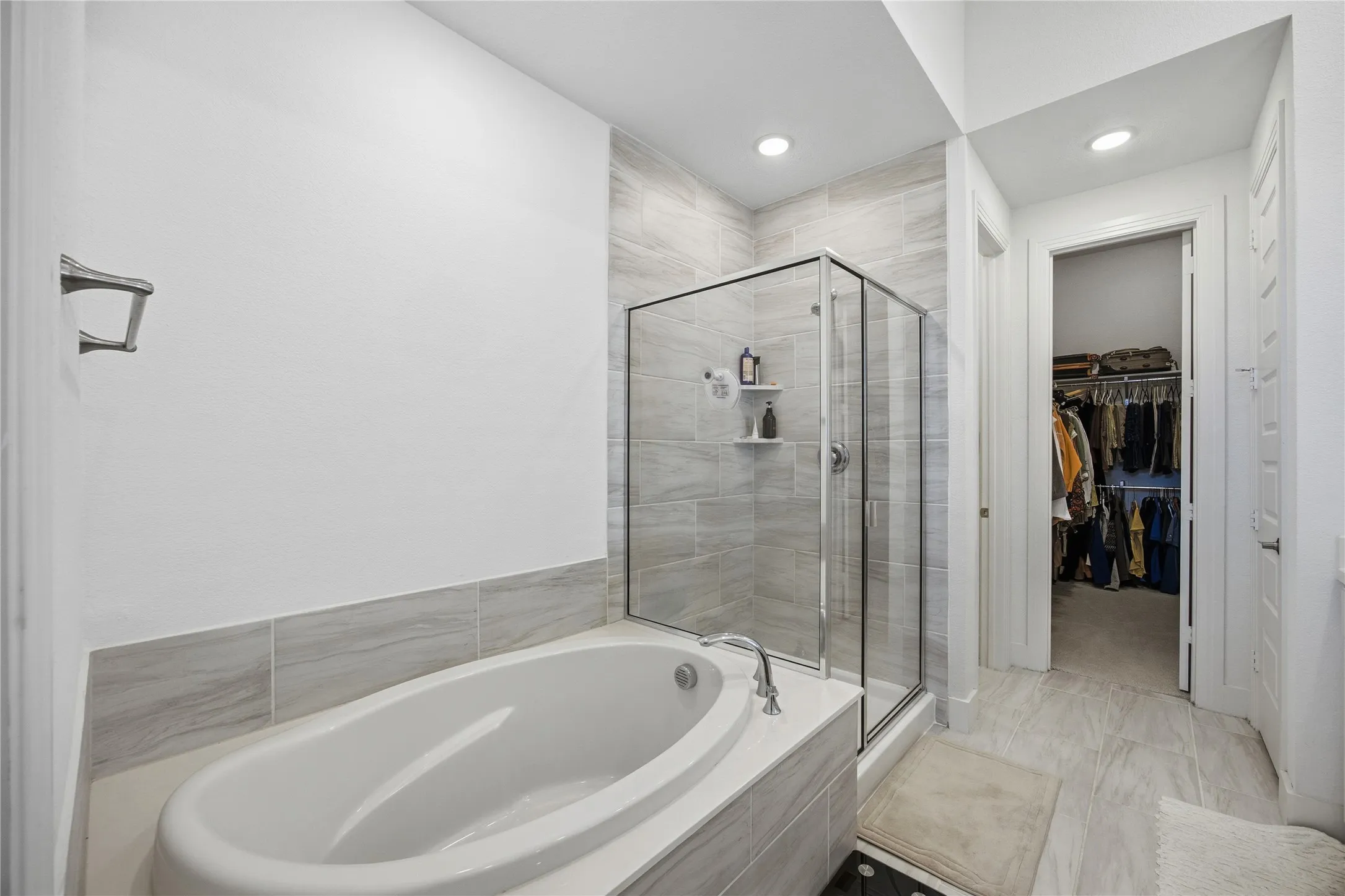 Freestanding soaking tub, glass-enclosed shower, tiled bathroom flooring, recessed lighting, and a walk-in closet with built-in organization
