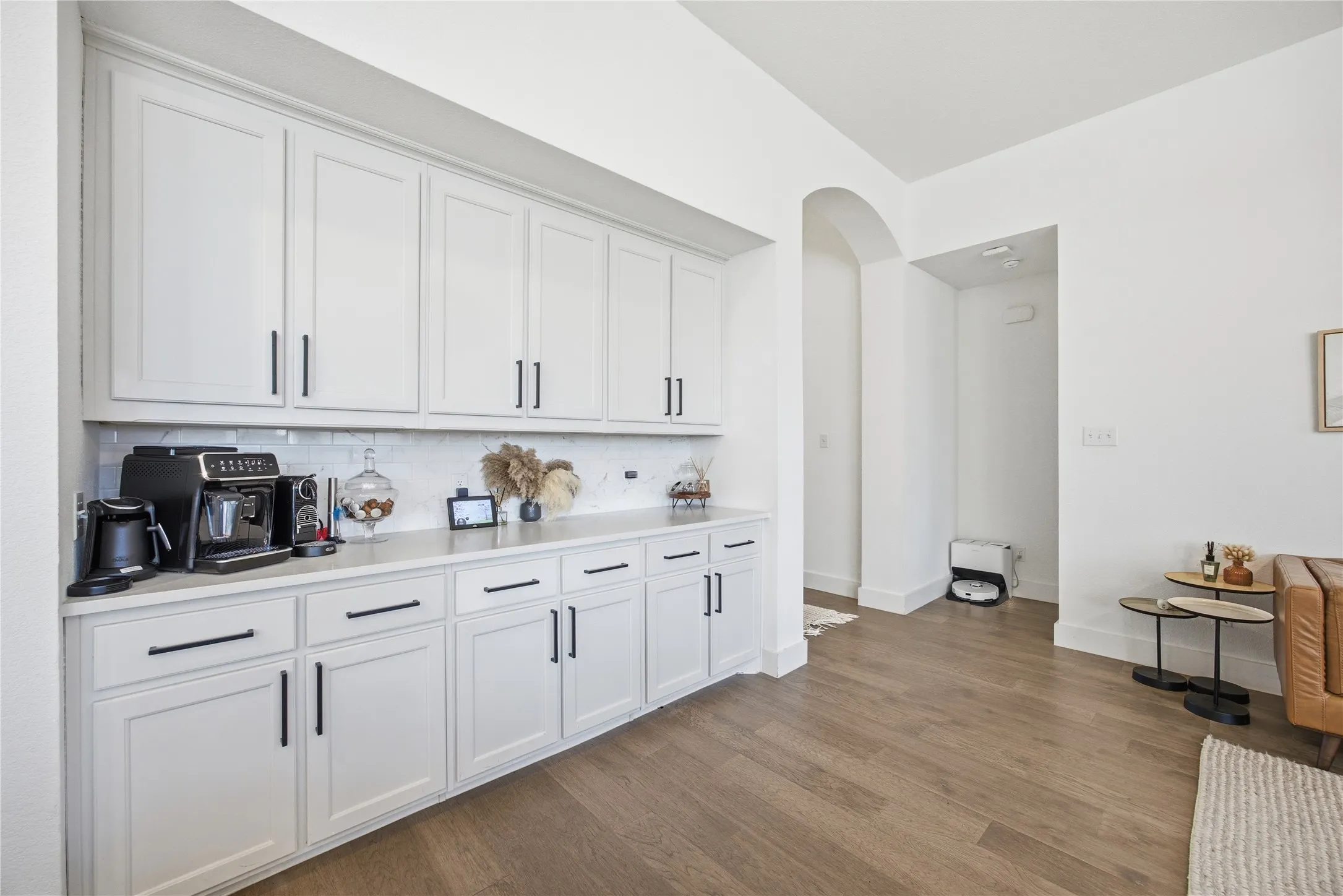 White cabinetry with matte black hardware, white subway tile backsplash, light-toned countertop, wood-finish flooring, and arched entryway