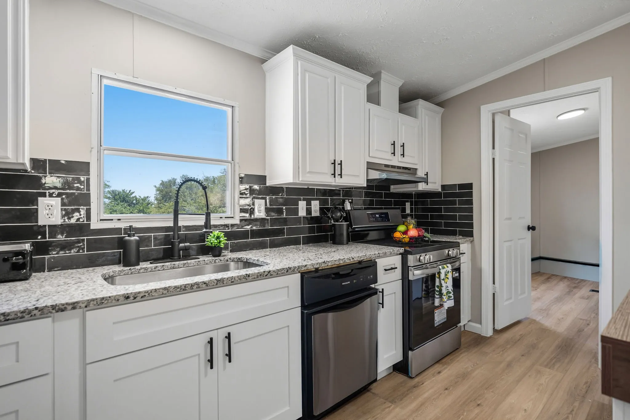 White shaker cabinetry, black subway tile backsplash, stainless steel sink, wood-finish flooring, and a window with natural light