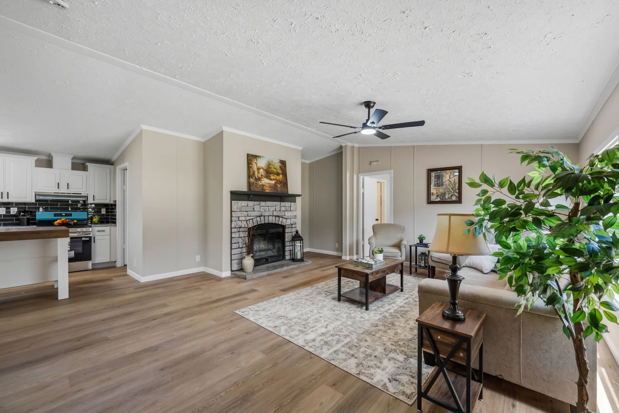 Open-concept living area featuring wood-finish flooring, a stone-surround fireplace with a dark mantel, a ceiling fan, and white cabinetry in the kitchen area
