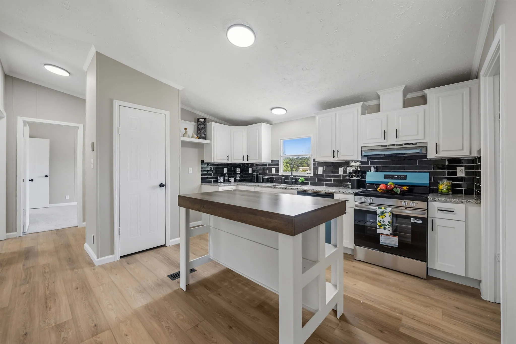Wood-finish flooring, white cabinetry with black hardware, dark subway tile backsplash, stainless steel appliances, and a central island with a wood-finish countertop