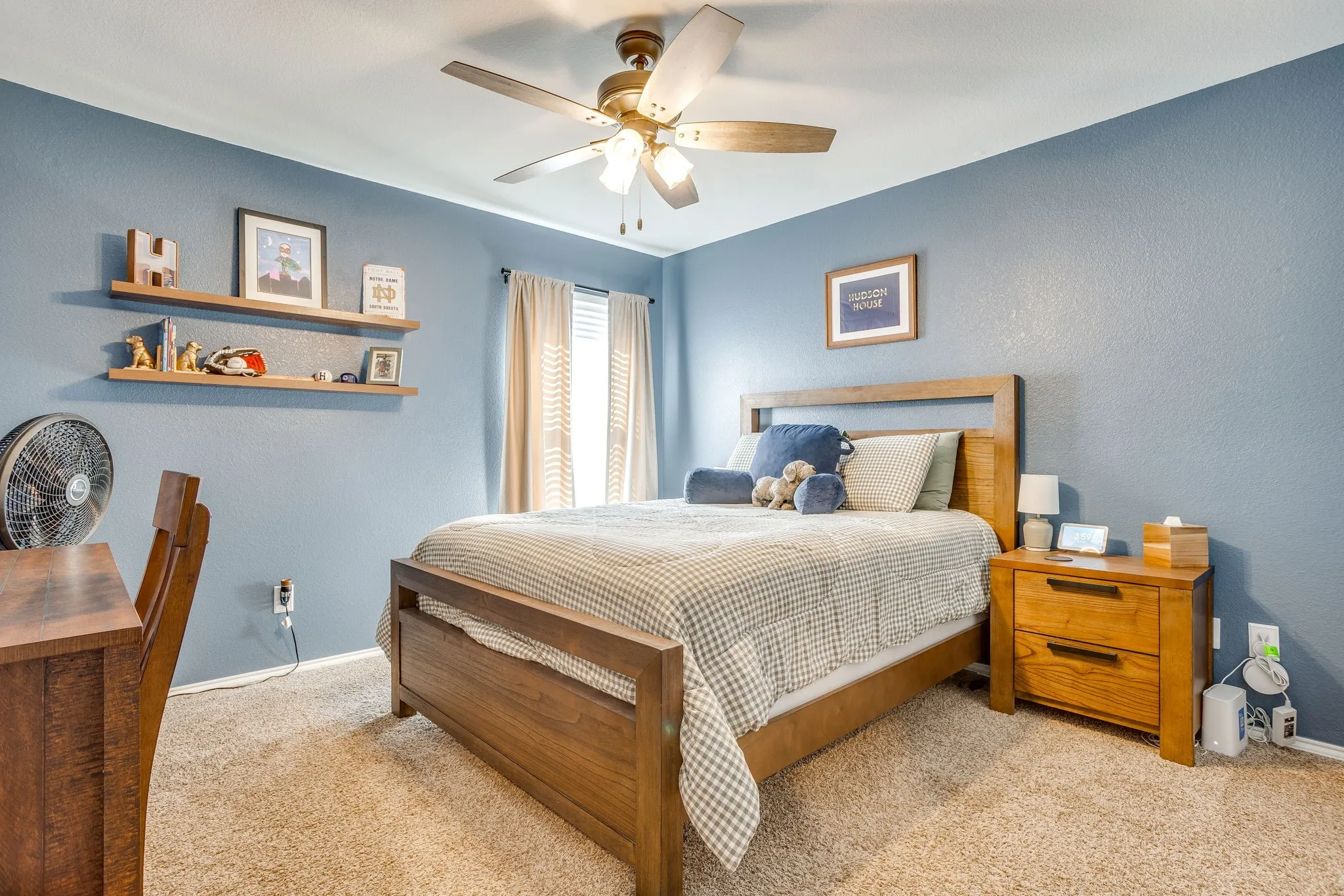 Ceiling fan with light fixture, carpeted flooring, window with drapes, floating wall shelves, and a wooden nightstand with two drawers