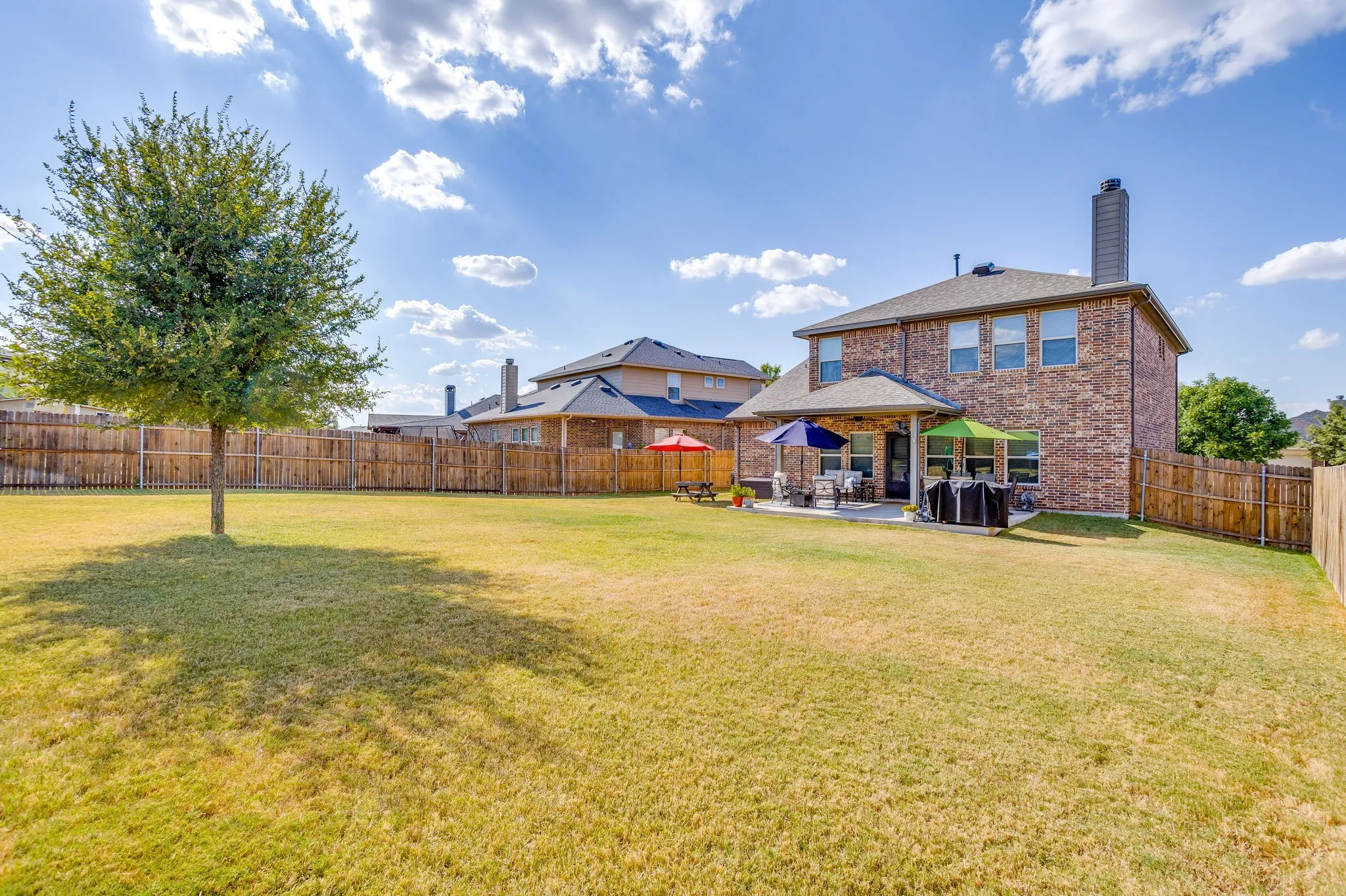 Expansive backyard with a manicured lawn, full privacy fencing, mature tree, and a covered patio area