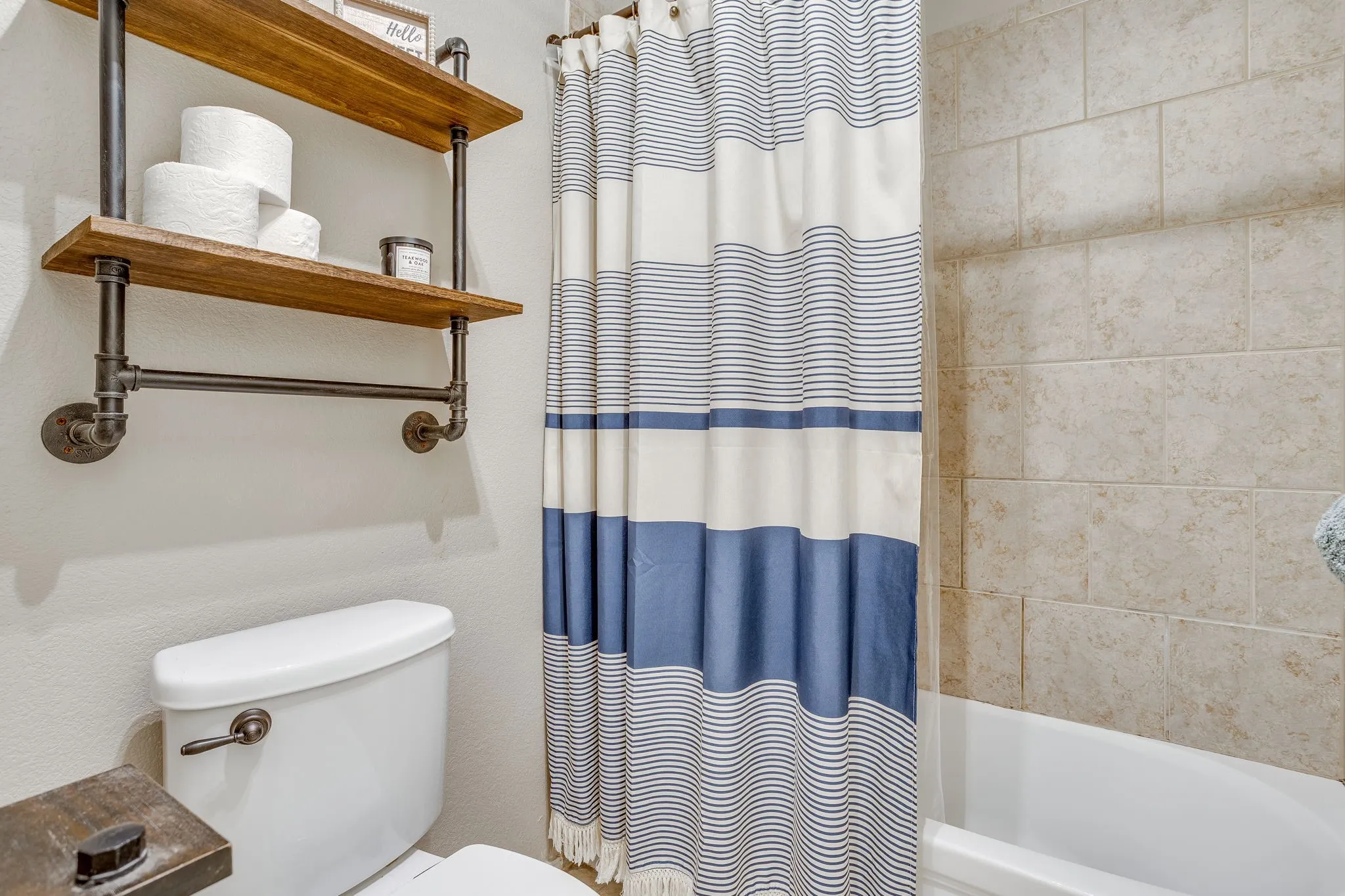 Tub-shower combination with light-toned rectangular wall tiles, white porcelain toilet, and industrial-style shelving with wood-finish shelves
