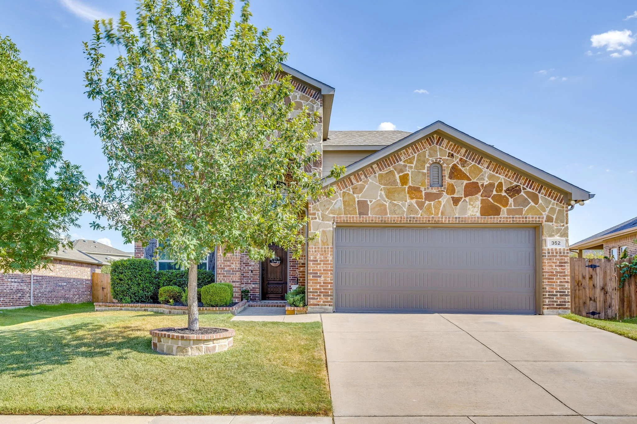 Brick and stone exterior featuring a two-car garage, mature landscaping, and an established tree
