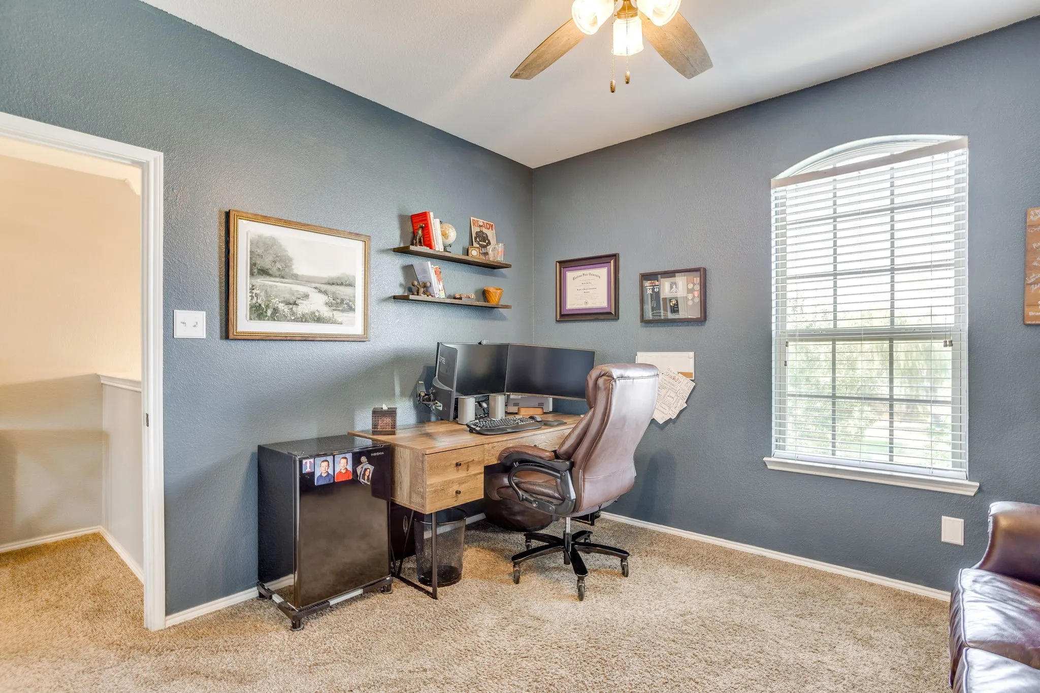 Arched window with blinds, carpeted flooring, two floating shelves, and a ceiling fan with integrated lighting