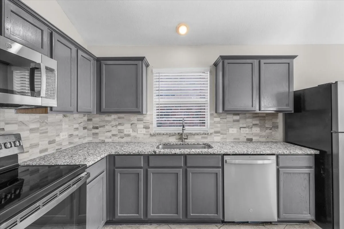 Kitchen featuring gray cabinetry, light-toned patterned tile backsplash, speckled countertops, stainless steel appliances, and a window with blinds