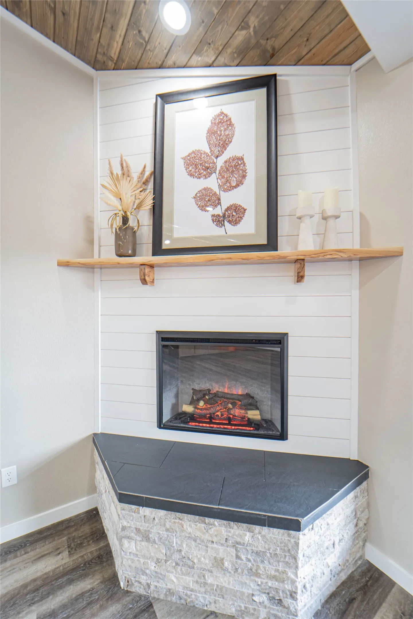 Corner electric fireplace featuring a stacked stone hearth, black tile top, shiplap surround, wood mantel, and wood-finish flooring