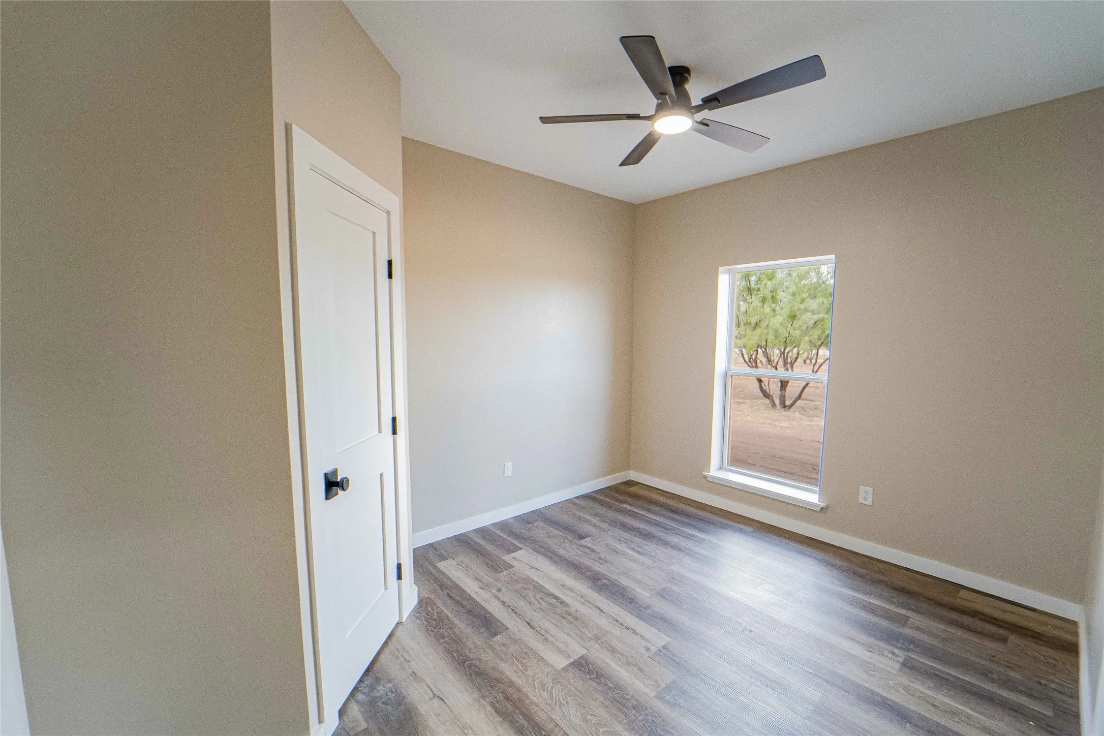 Wood-finish flooring, single window, white trim, flat-panel door, and a ceiling fan with integrated lighting