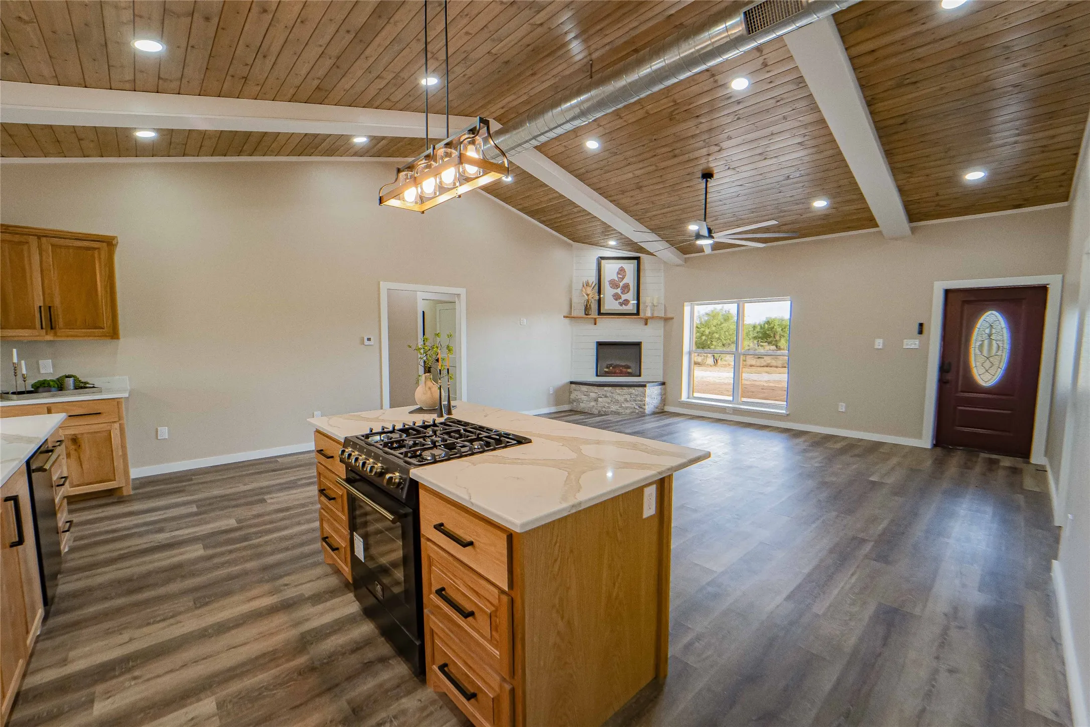 Vaulted wood-paneled ceiling with exposed beams, wood-finish flooring, kitchen island with built-in range, stone fireplace, and a decorative front entry door