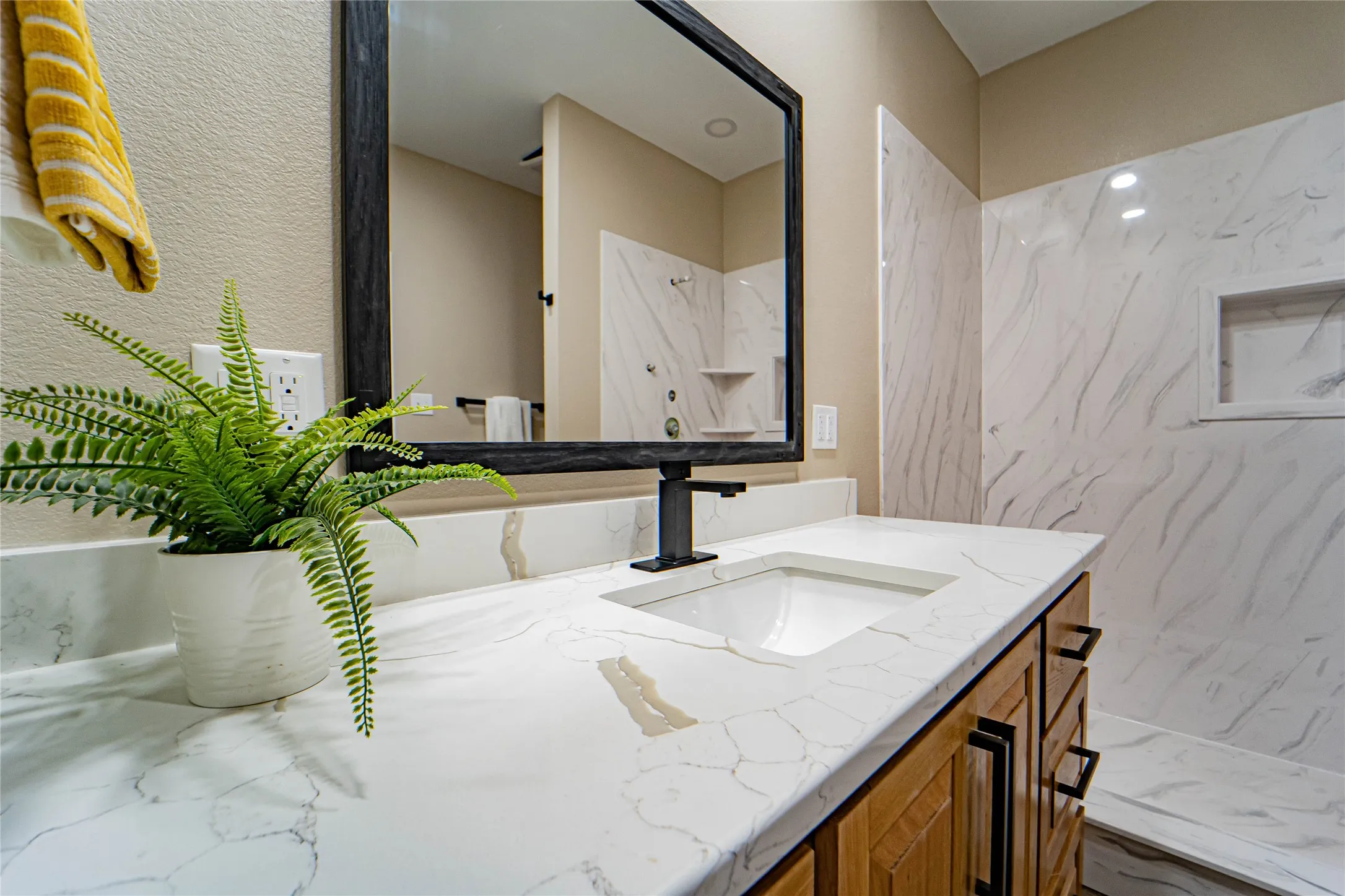 Integrated vanity with an undermount sink, a dark-framed mirror, wood-finish cabinetry with dark hardware, a single-handle dark faucet, and a shower with white and grey patterned wall tile