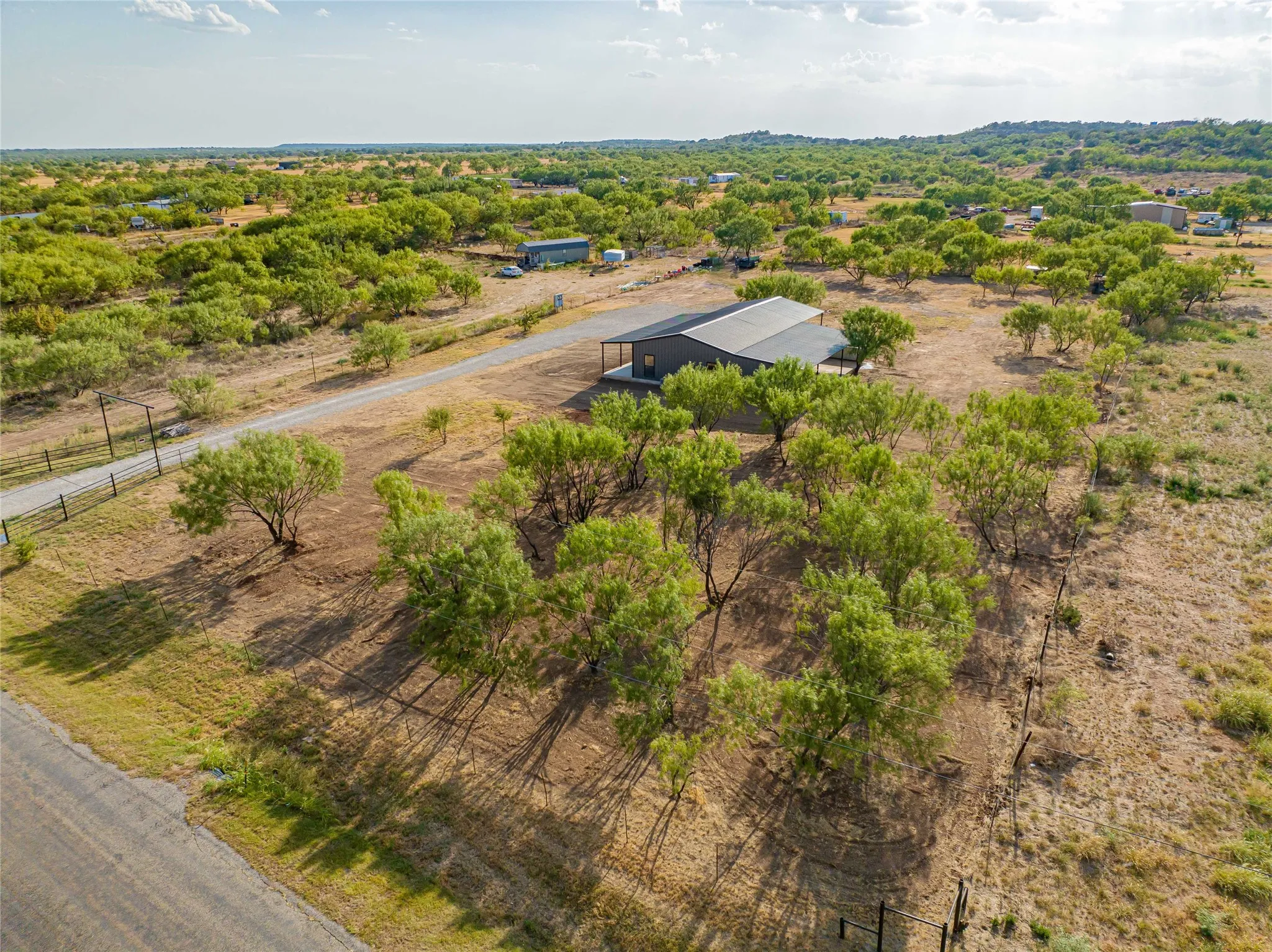 Expansive rural property featuring a modern structure with a metal roof, surrounded by numerous mature trees, and a paved access road