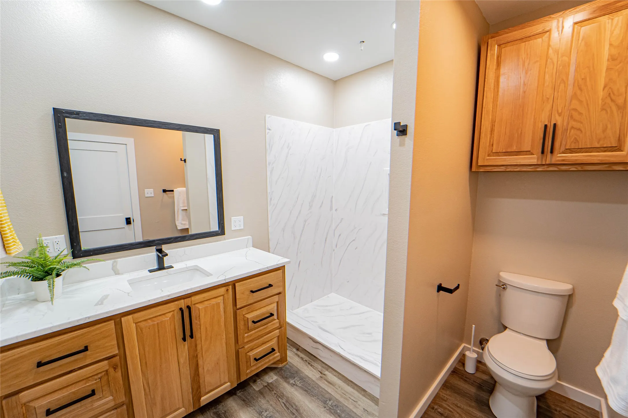 Vanity with quartz-finish countertop and undermount sink, wood-finish cabinetry, walk-in shower with textured wall panels, and wood-finish flooring