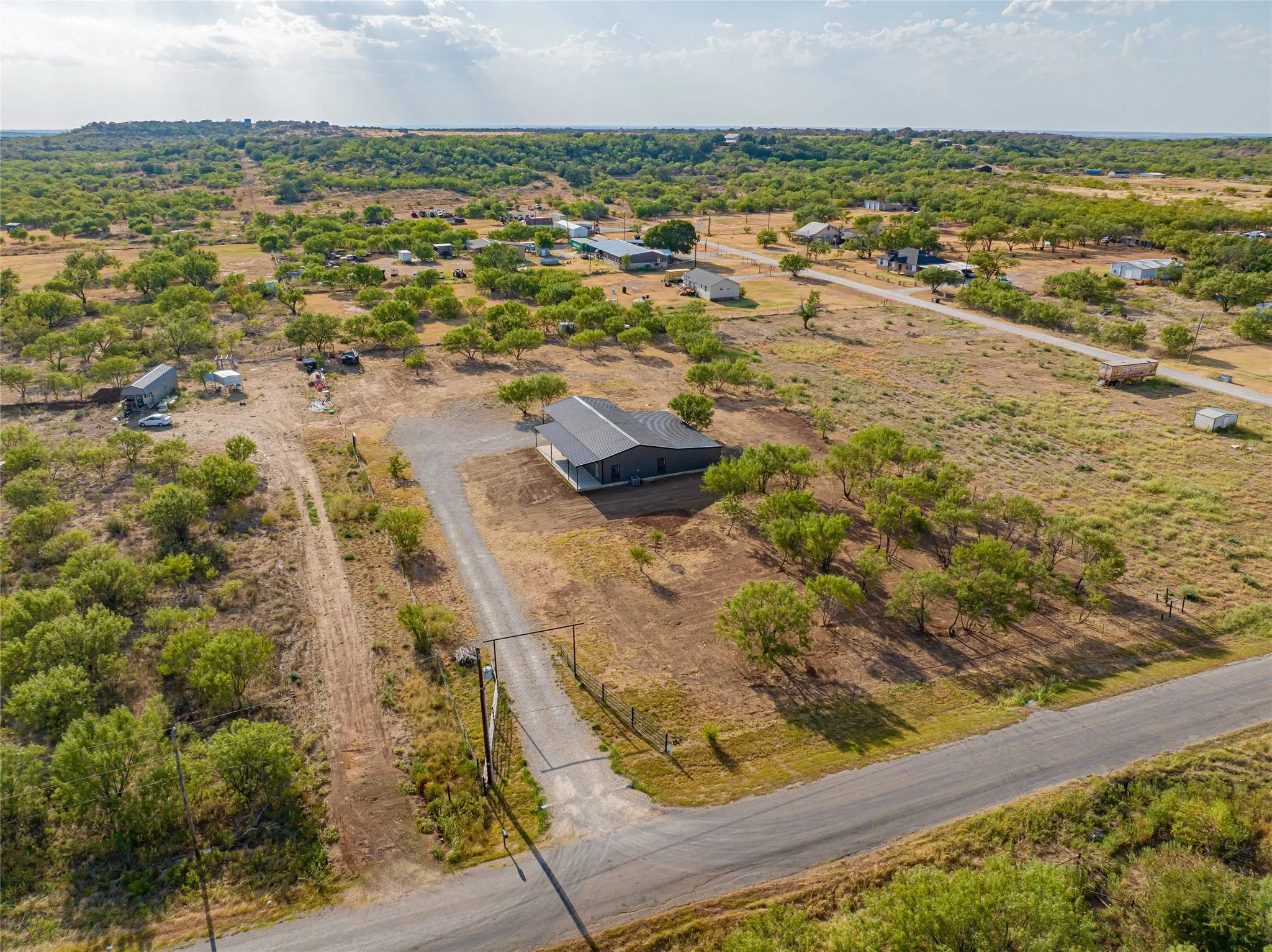 Expansive land with natural vegetation, a paved road, and a gravel driveway leading to a residence with a dark-colored exterior and a sloped roof
