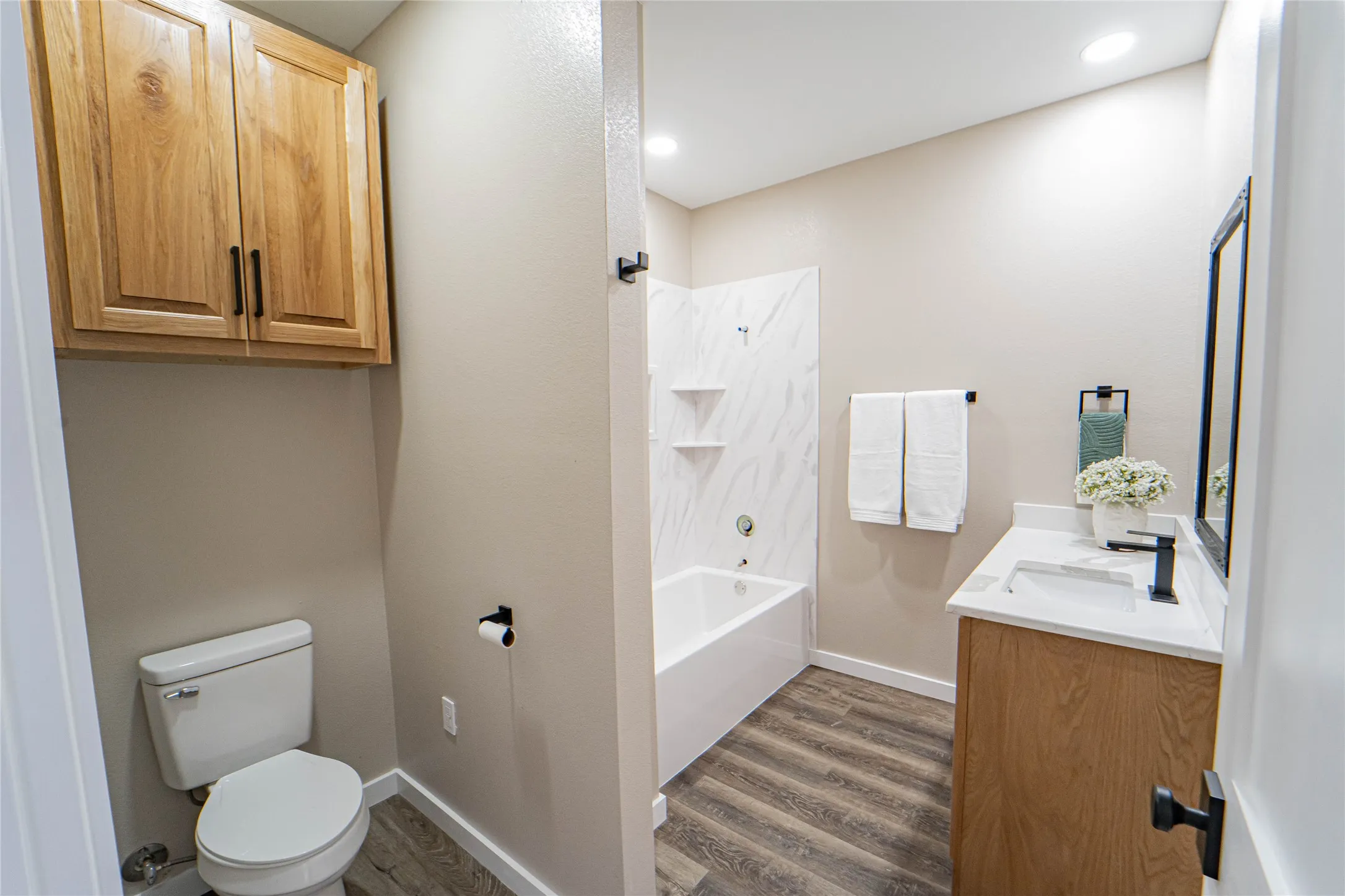 Wood-finish flooring, light wood cabinetry, white countertop with integrated sink, black-framed mirror, and white tiled tub surround