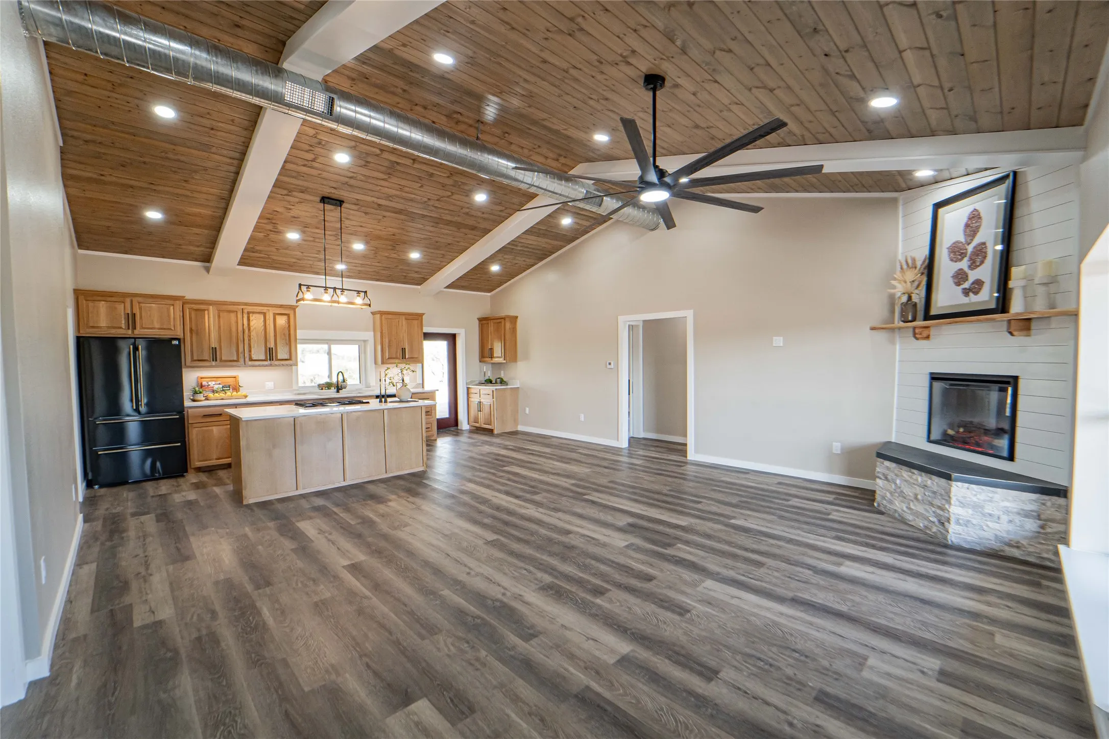 Vaulted wood-plank ceiling with exposed ductwork, wood-finish flooring, kitchen island with sink, shaker-style cabinetry, and a corner fireplace with stone-clad hearth