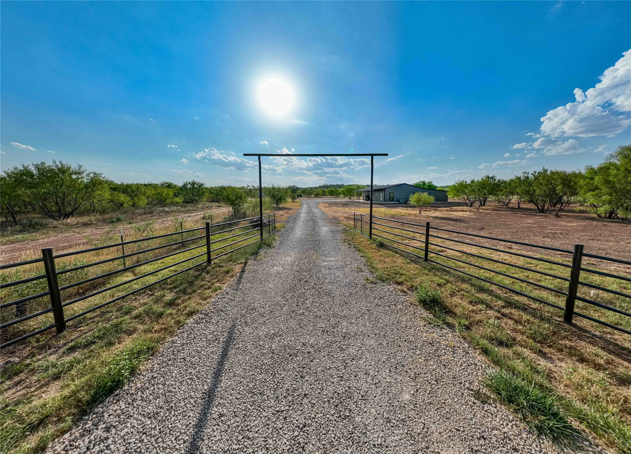 Gravel driveway framed by metal fencing, leading to a single-story structure with a metal roof