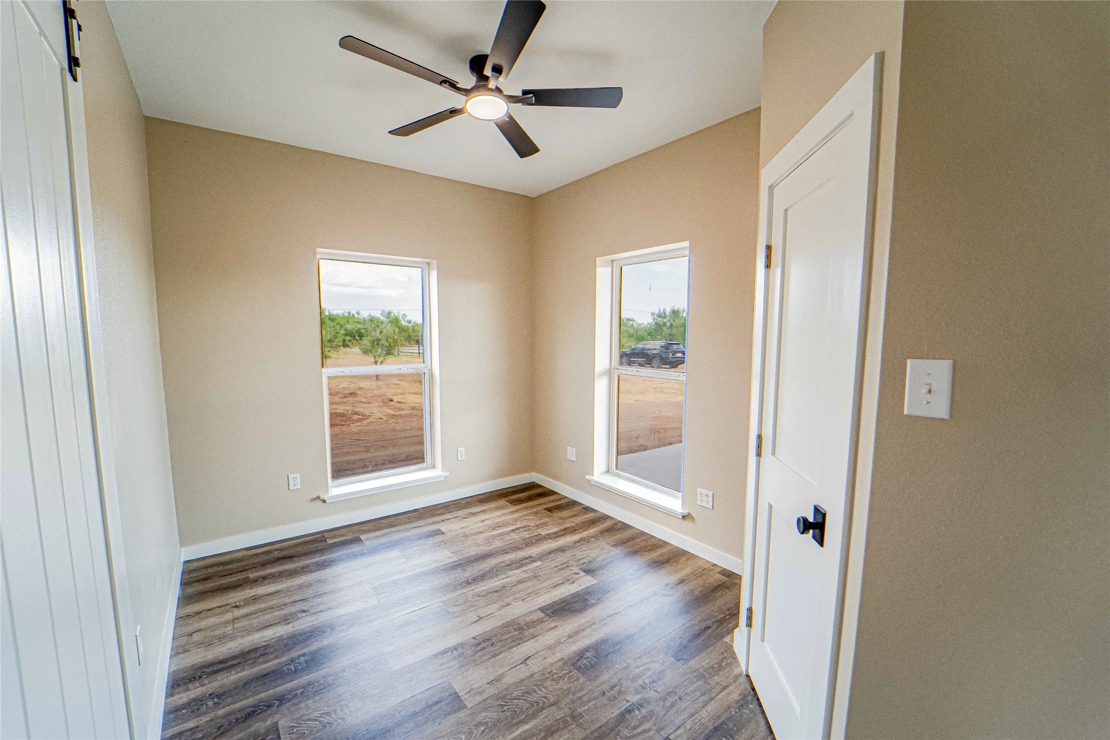 Wood-finish flooring, two windows with white trim, ceiling fan with light, and a white paneled door with black hardware