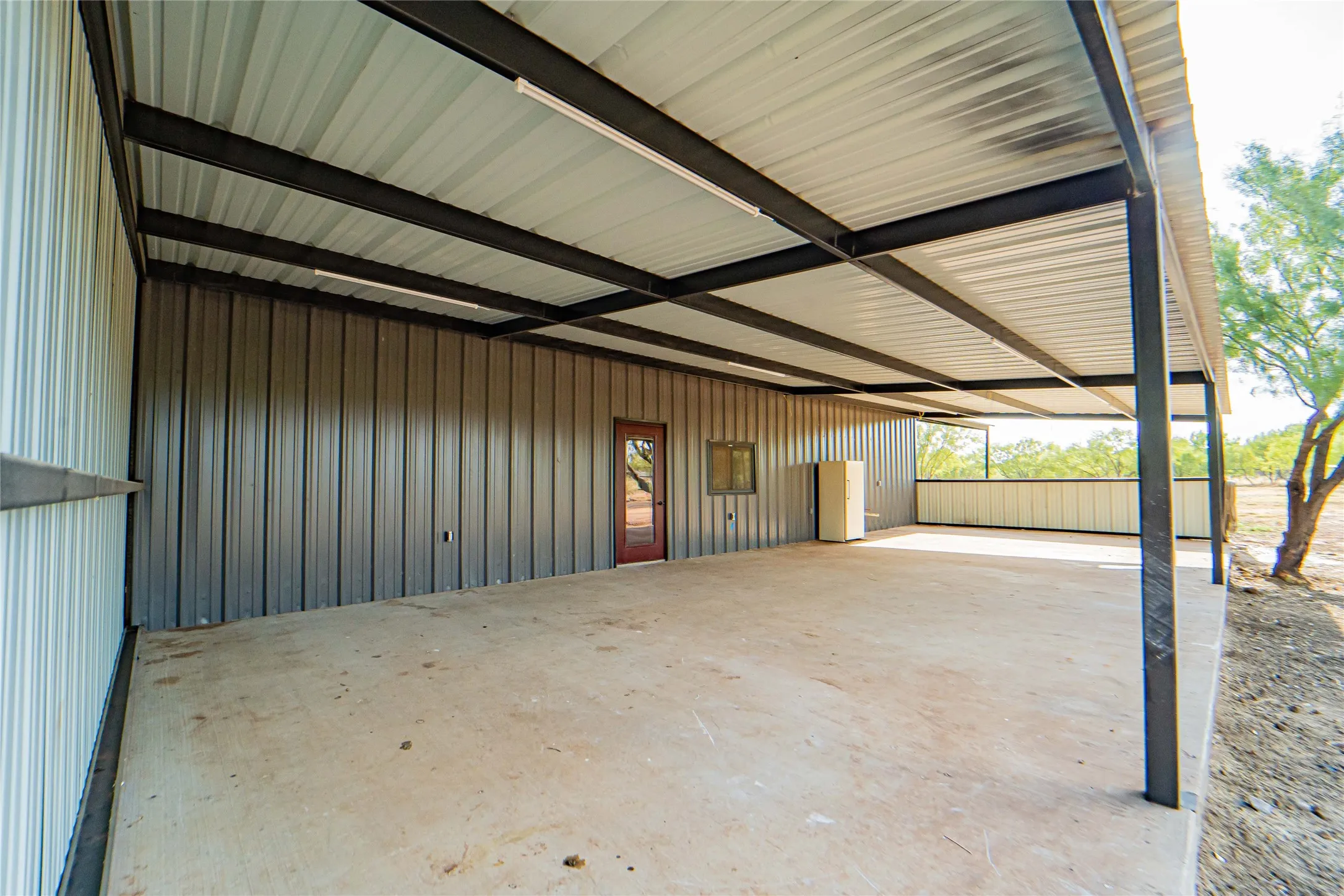 Covered concrete patio with metal support beams, metal siding, and a red-stained door