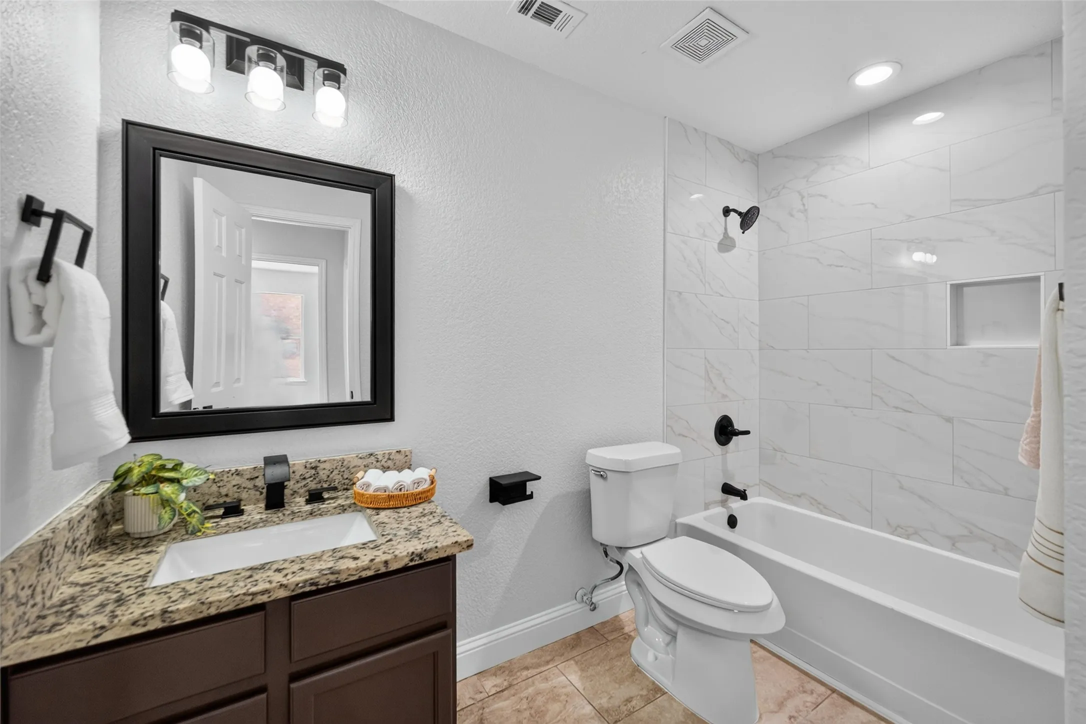 Bathroom featuring a built-in tub with a shower, white tile surround, dark wood-finish vanity with a light-toned countertop, and a rectangular undermount sink with a matte black faucet