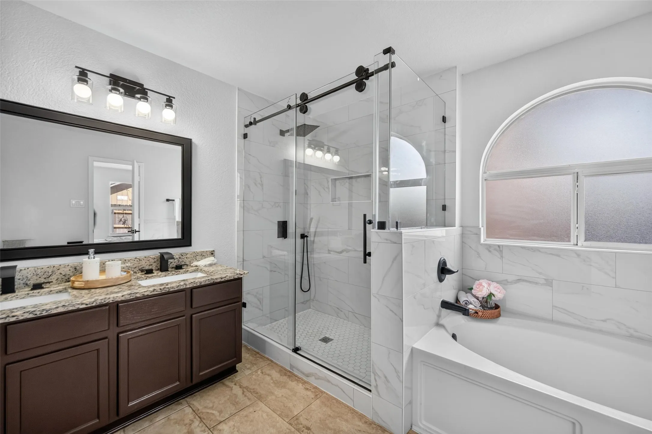 Dual sink vanity with granite countertop, large framed mirror, glass-enclosed shower with sliding barn door, and a built-in tub with tile surround