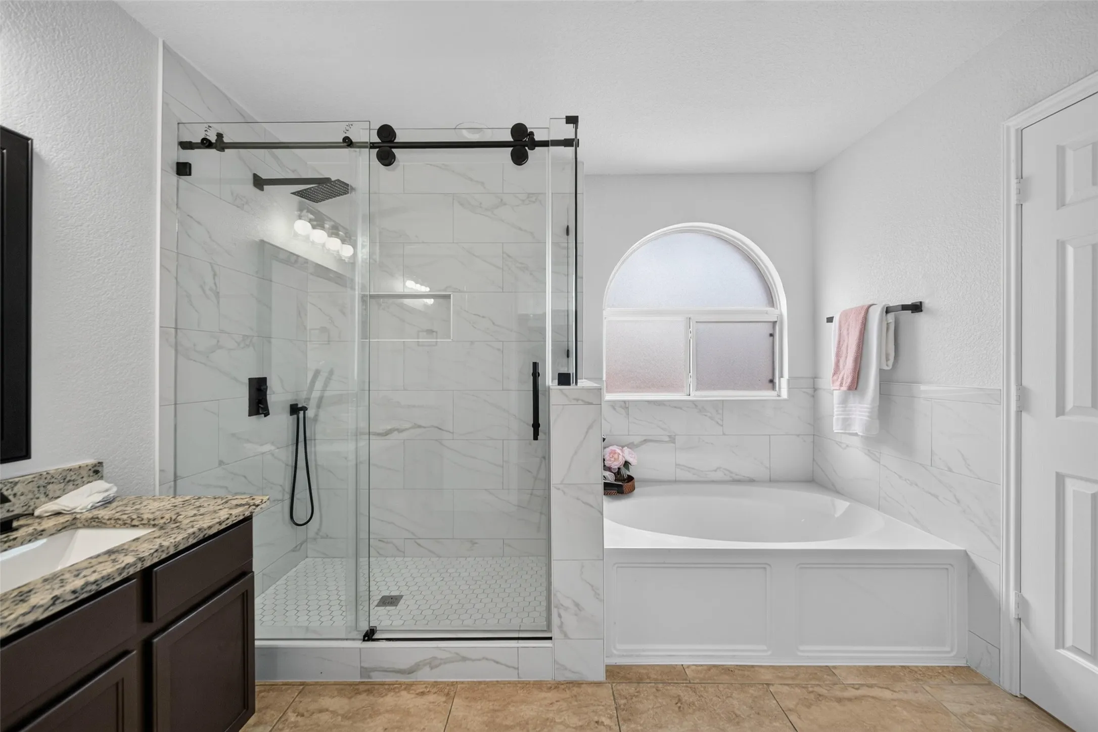 Walk-in shower with clear glass enclosure and black hardware, freestanding soaking tub, arched frosted window, single vanity with light-colored countertop, and large format floor tile