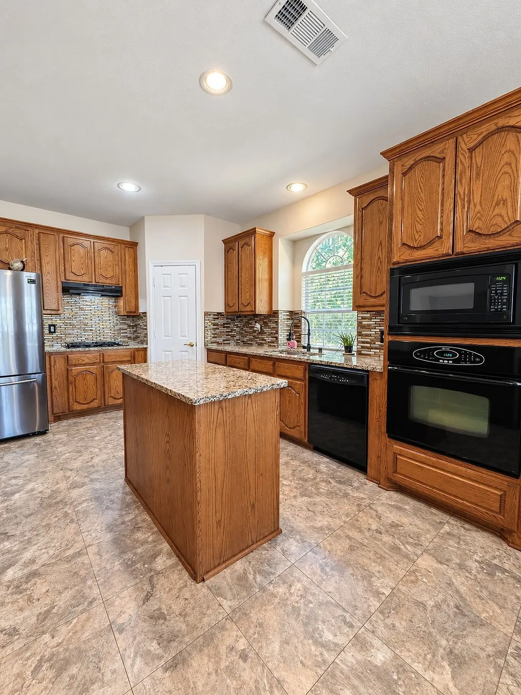 Kitchen featuring wood cabinetry, a center island with a stone-finish countertop, black appliances, and a tiled backsplash