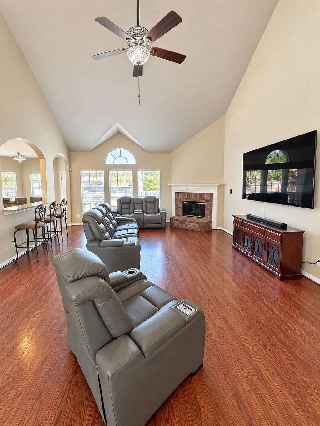Vaulted ceiling, wood-finish flooring, brick-faced fireplace, ceiling fan with integrated lighting, and arched window