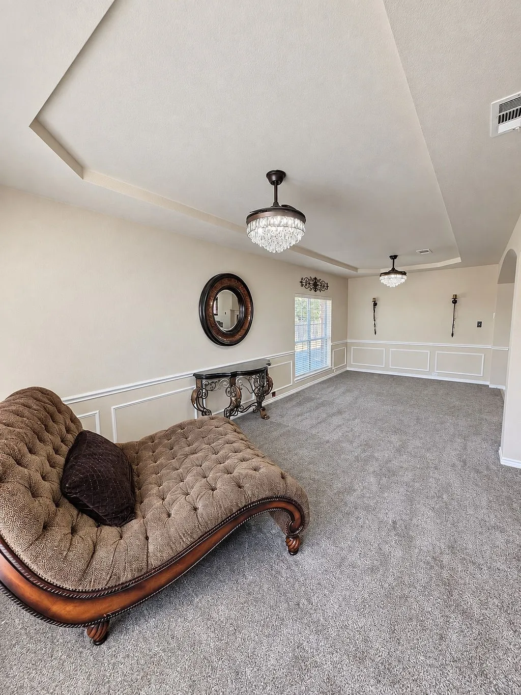 Tray ceiling with decorative trim, multiple crystal-style ceiling light fixtures, wainscoting, arched doorway, and neutral tone carpeting