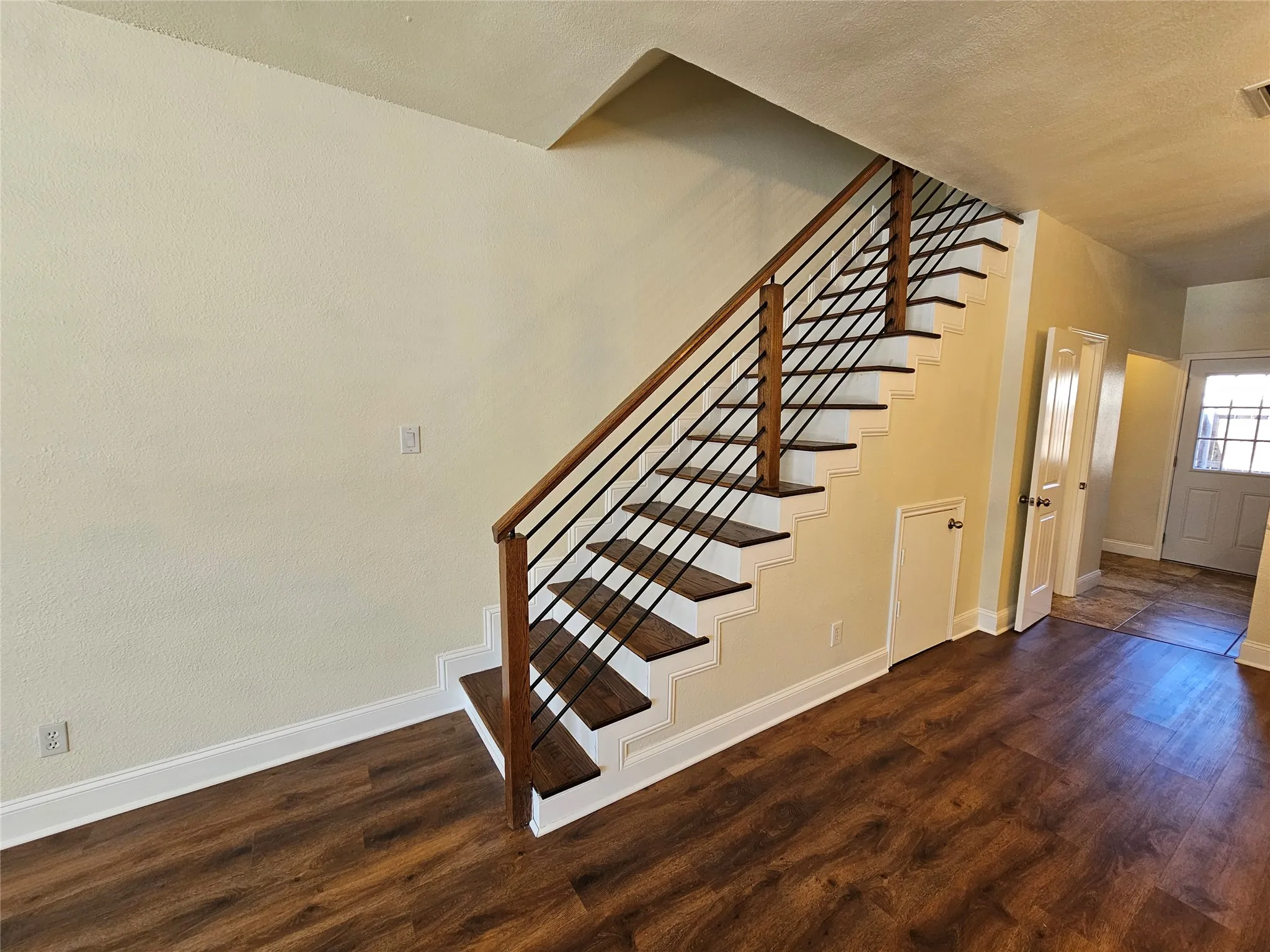 Wood-finish flooring, staircase with dark wood treads, metal balusters, and wood handrail, white trim, and a white entrance door with decorative glass
