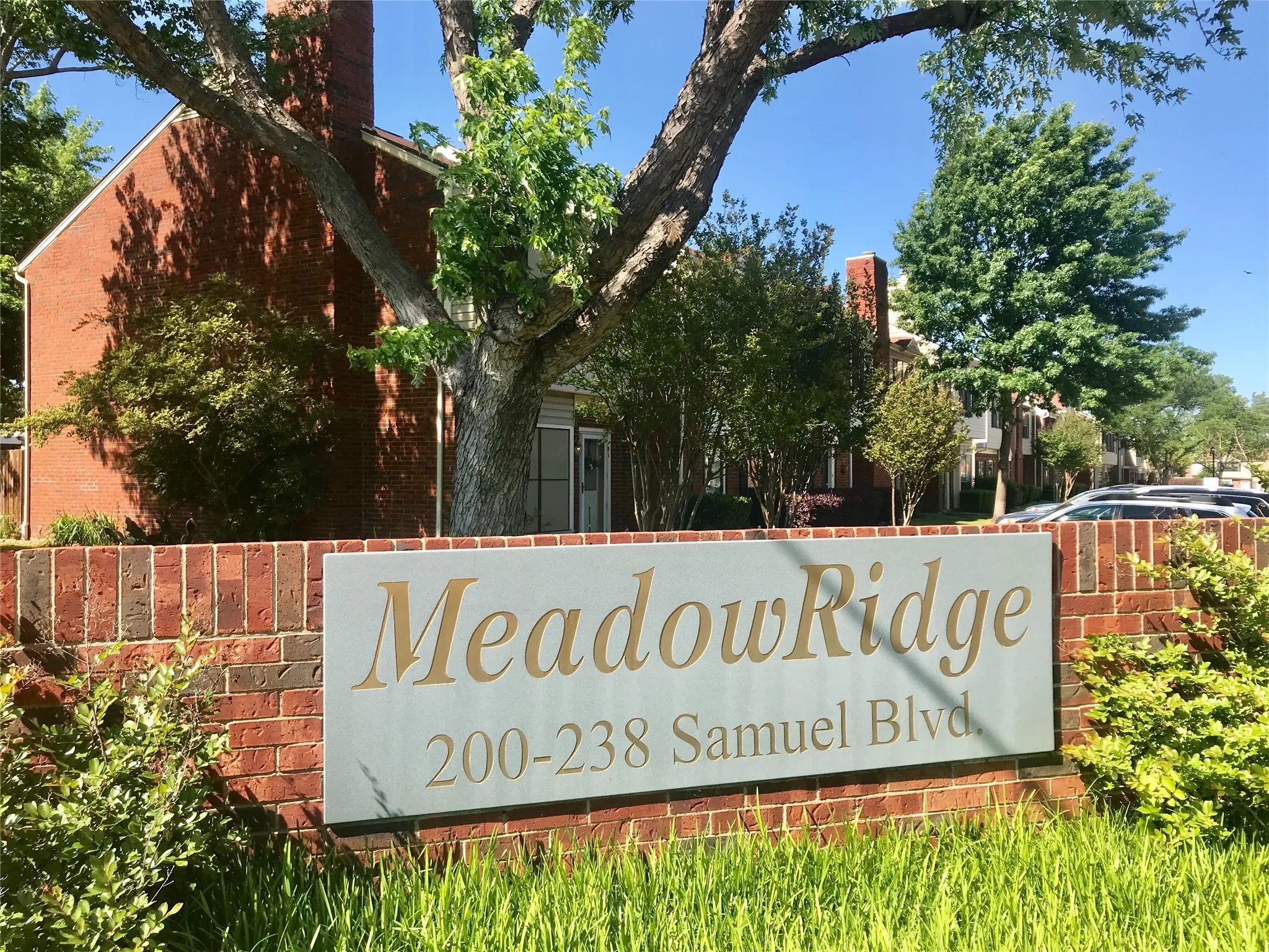 Brick entryway wall with a prominent community sign, featuring established landscaping and mature trees