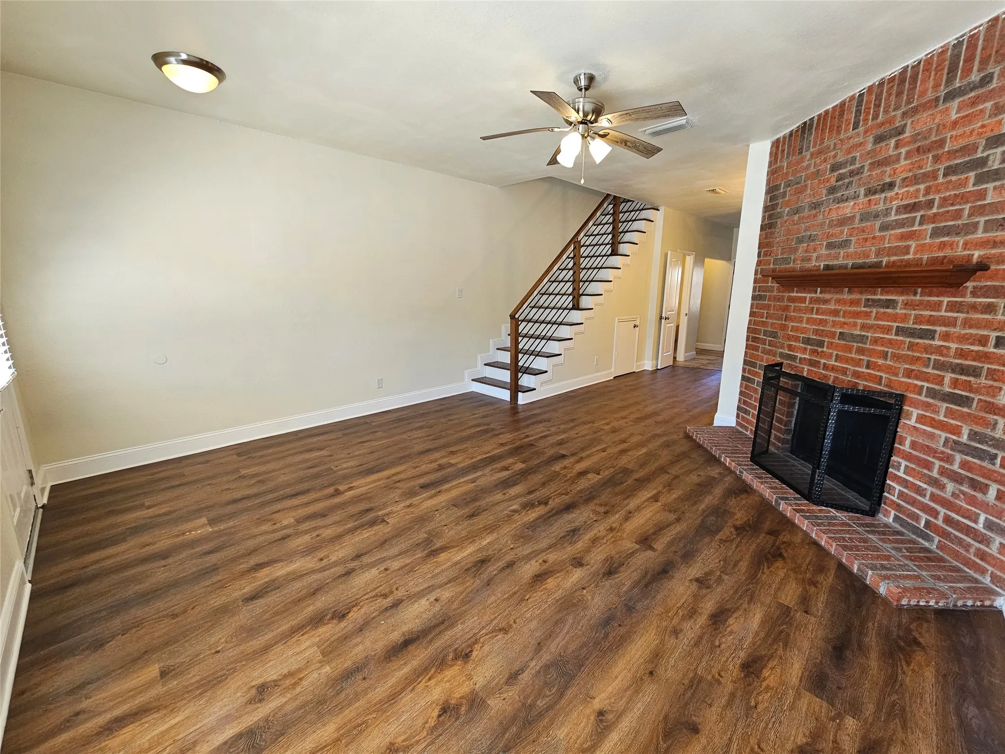 Wood-finish flooring throughout, brick fireplace with a wood mantle, ceiling fan, and staircase with metal balusters