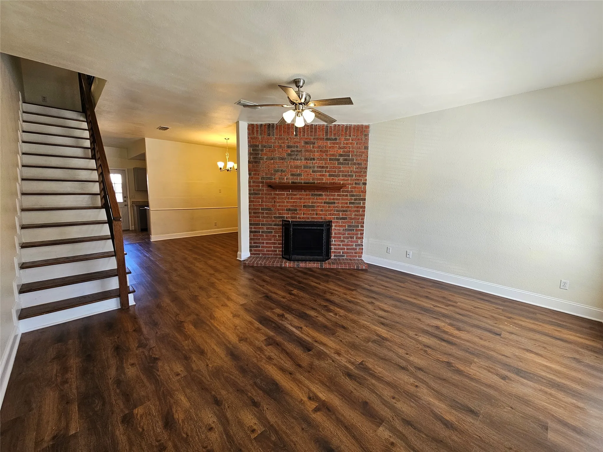Wood-finish flooring throughout, featuring a brick fireplace with a wood mantel, an integrated ceiling fan with lighting, and a staircase with wood railing