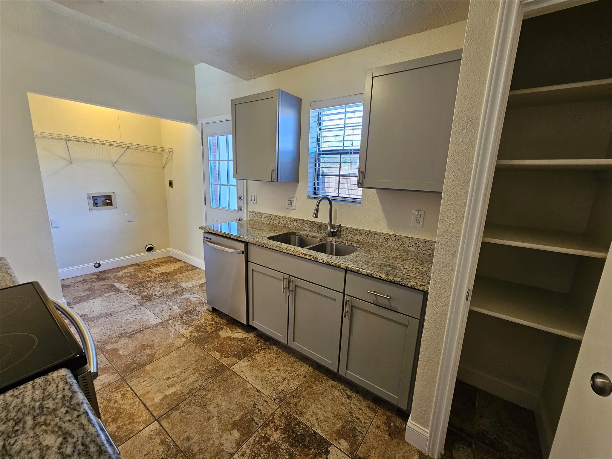 Granite-style countertops, stainless-steel dishwasher, gray shaker-style cabinetry, a built-in pantry with shelving, and multi-toned tile flooring