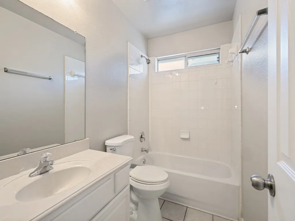 White vanity with integrated sink, chrome faucet, white tile flooring, white tile shower surround, and a white bathtub