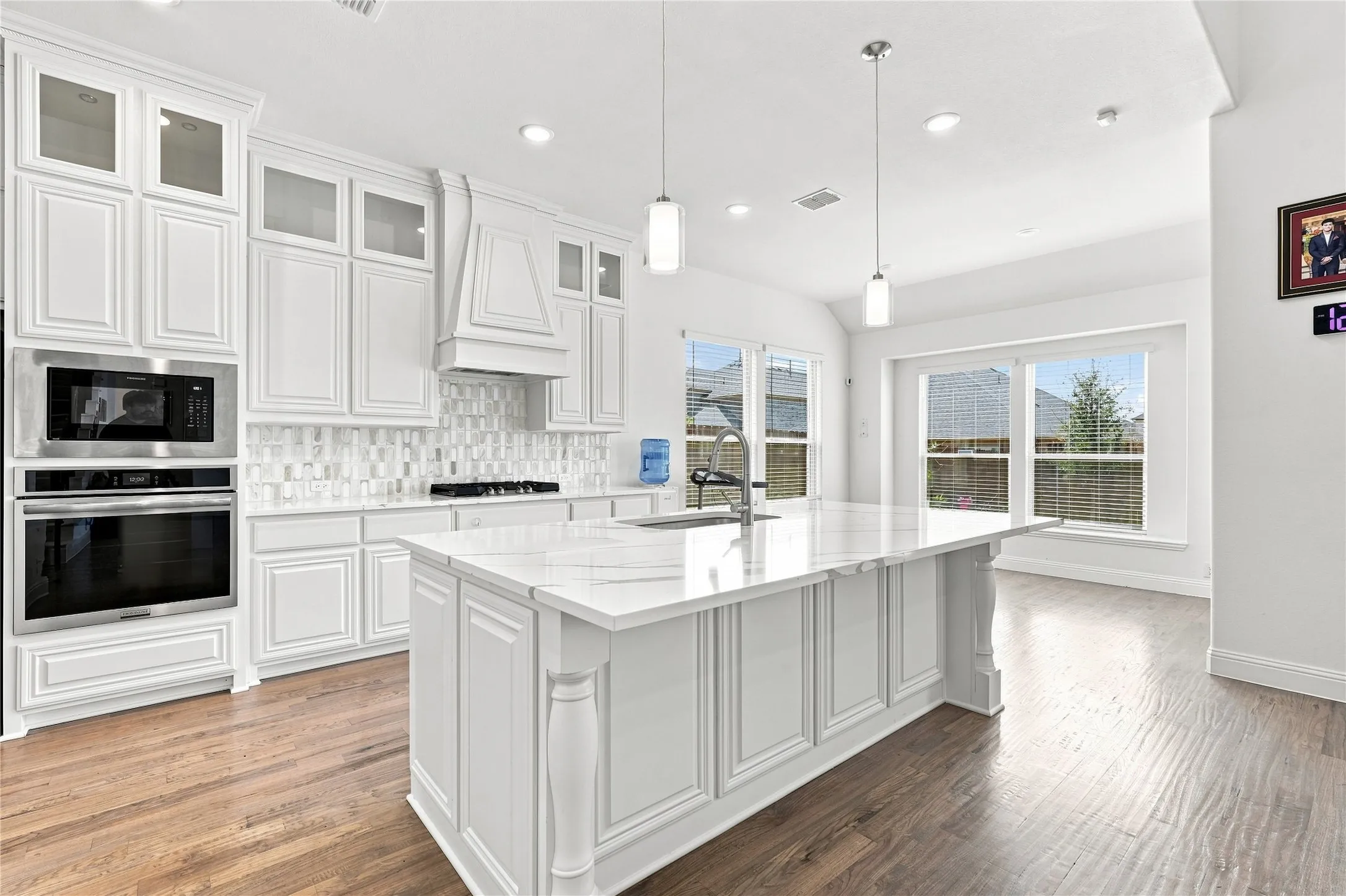 White cabinetry with glass-front accents, built-in stainless steel appliances, a large center island with a sink, a decorative tile backsplash, and wood-finish flooring