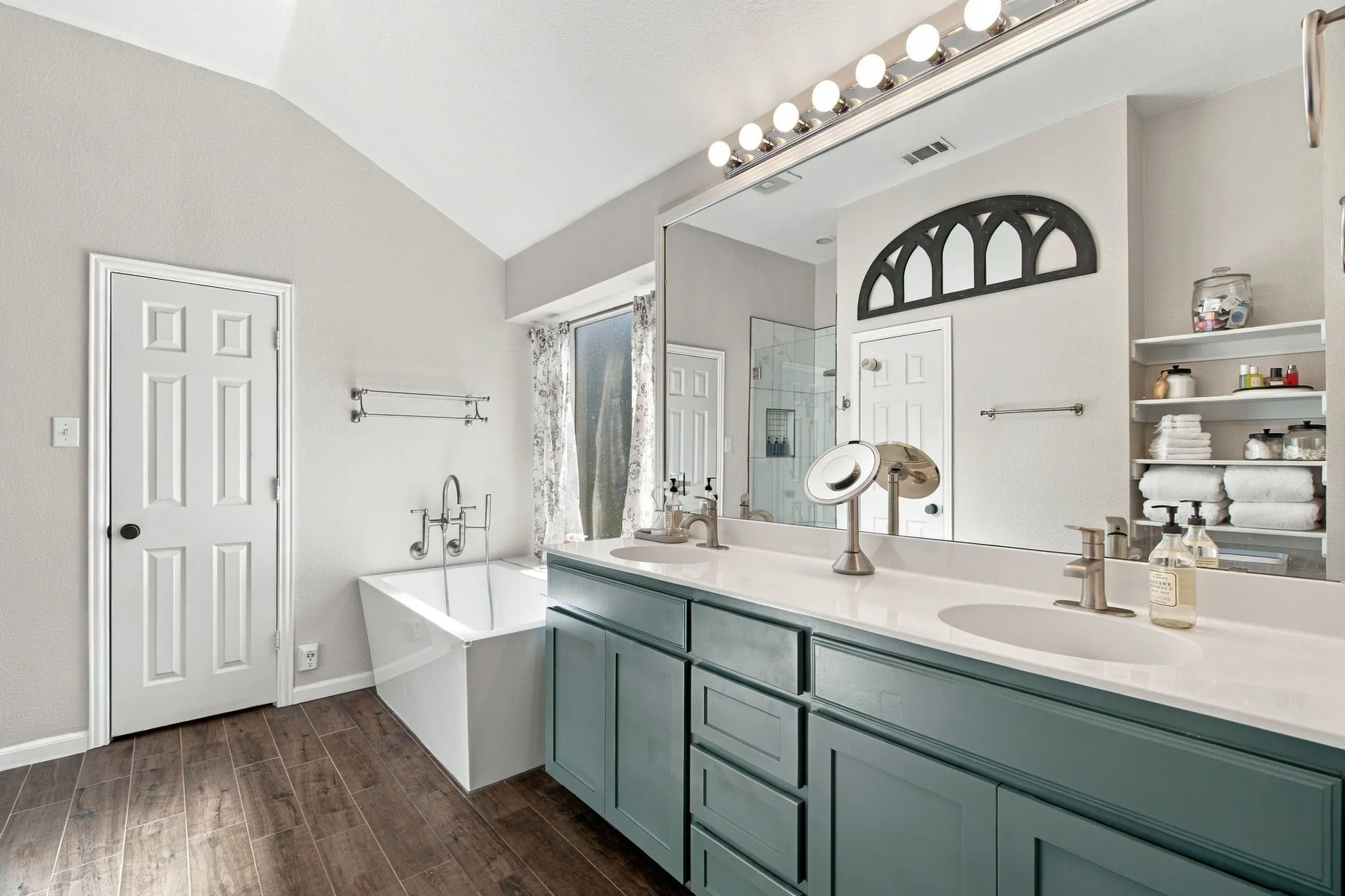 Spacious bathroom featuring a double vanity with white countertops, a freestanding tub with wall-mounted faucet, and wood-finish flooring
