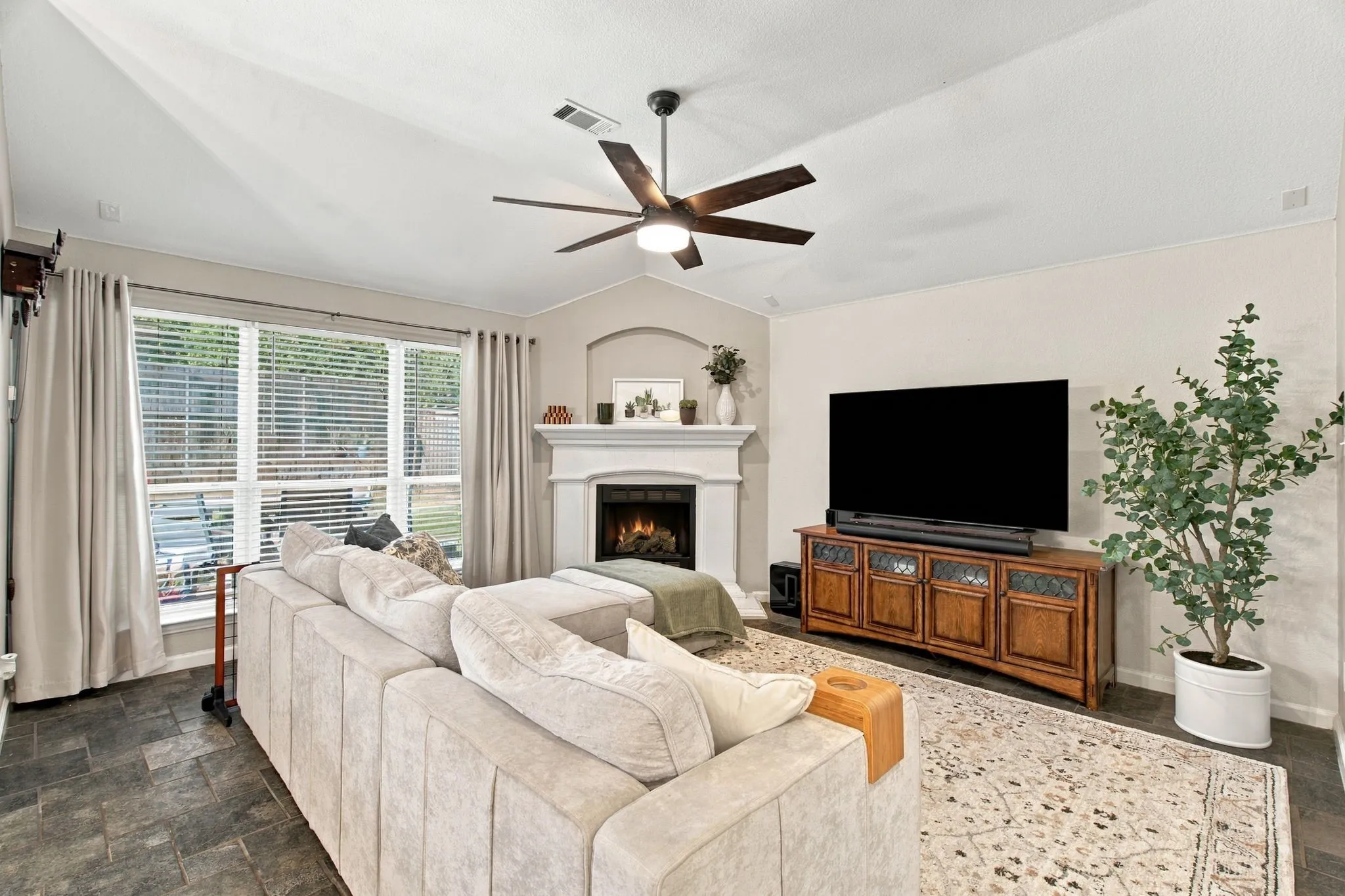 Vaulted ceiling with a ceiling fan, fireplace with a mantle, large window with blinds, and tile flooring