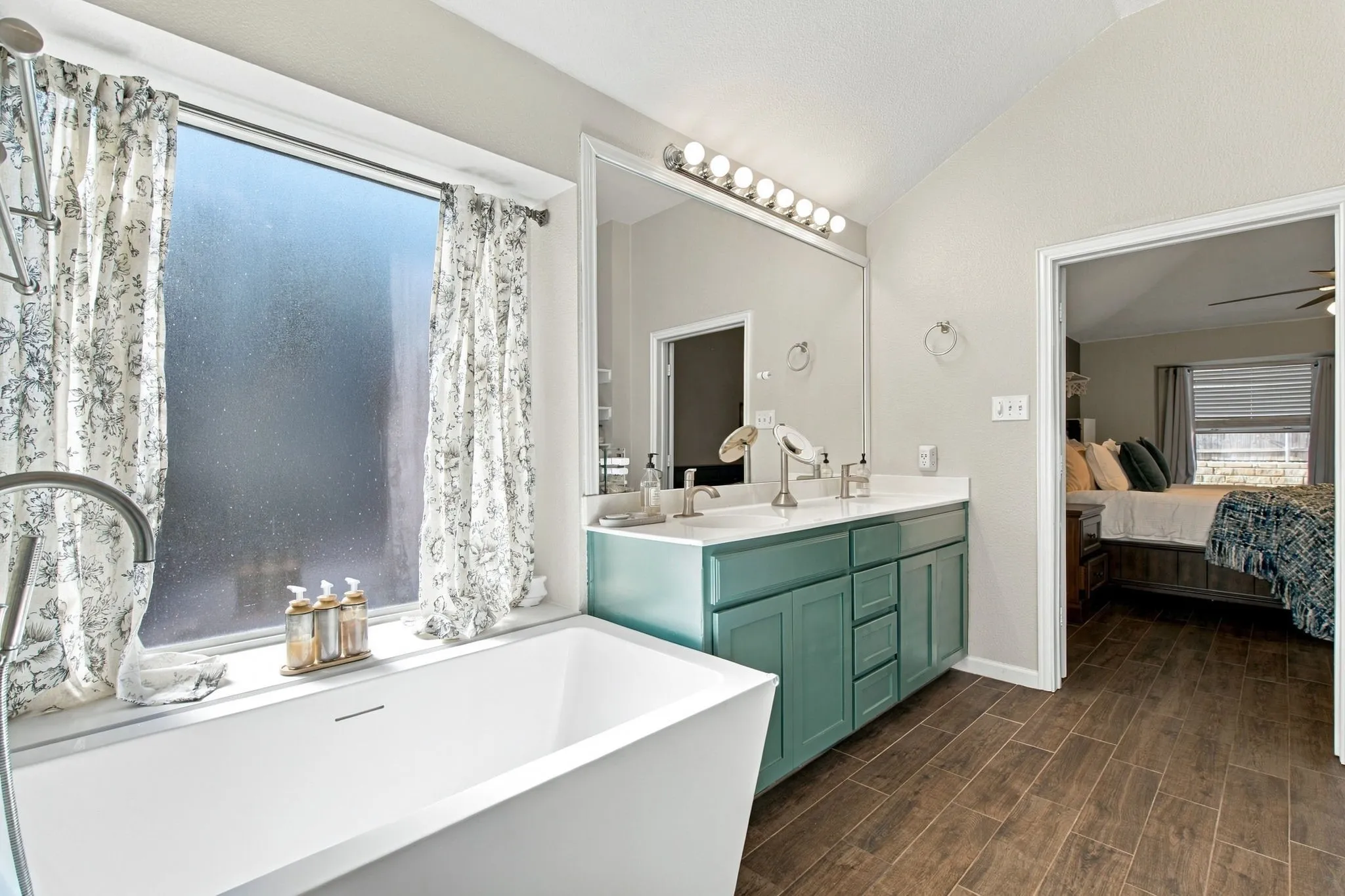 Soaking tub with a wall-mounted faucet, wood-finish flooring, dual vanity with cabinet storage, and a large mirror with overhead lighting