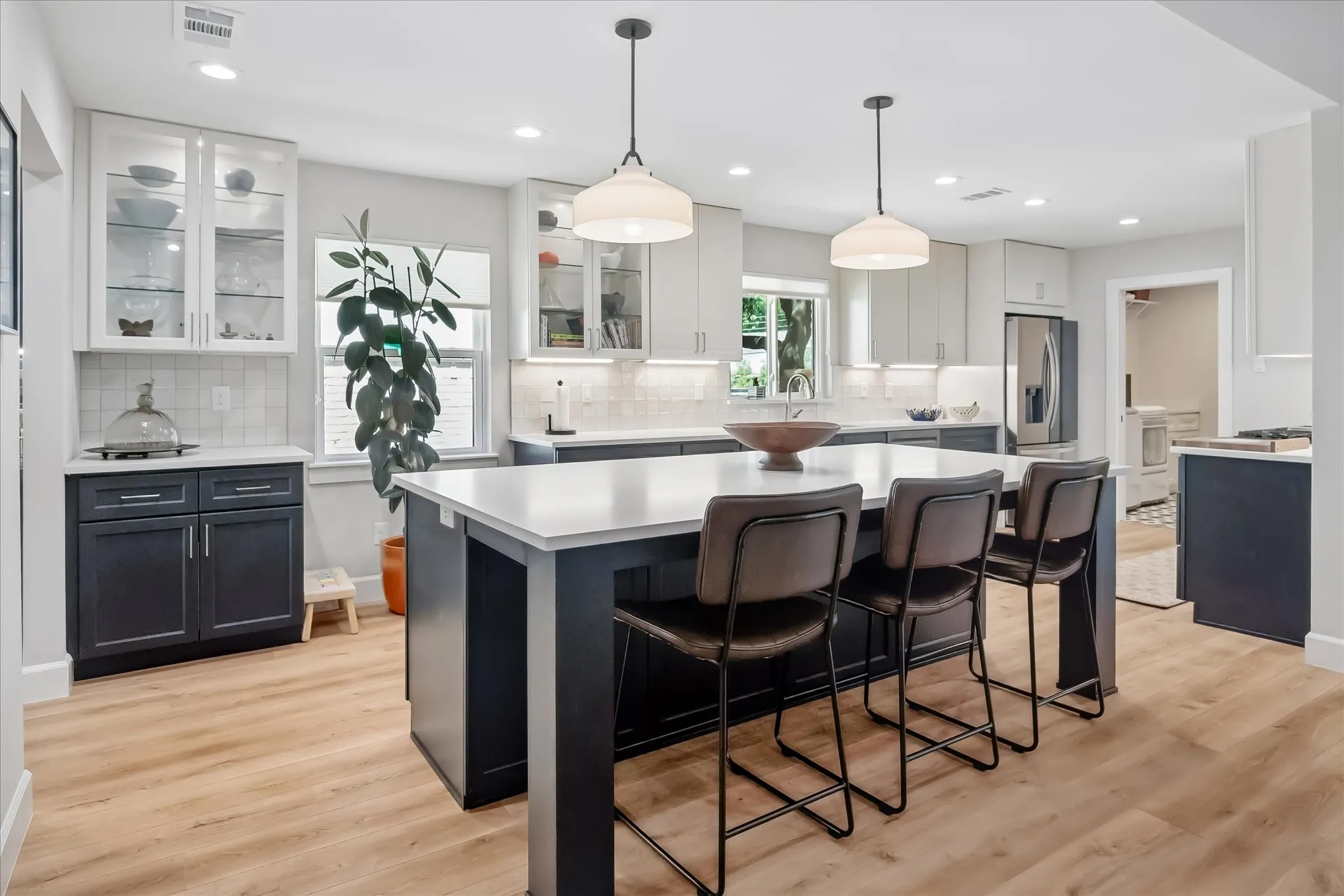 Kitchen featuring a large island with seating, pendant lighting, wood-finish flooring, white upper cabinetry with glass inserts, and dark lower cabinetry