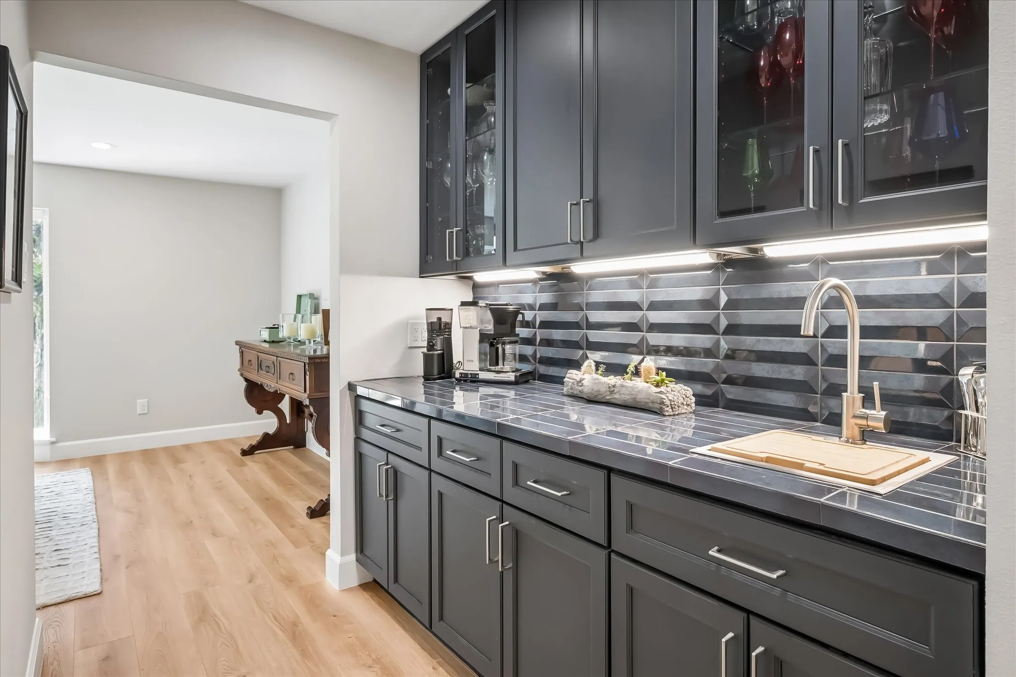 Built-in cabinetry with glass-front uppers, under-cabinet lighting, a textured tile backsplash, a stainless steel sink, and wood-finish flooring