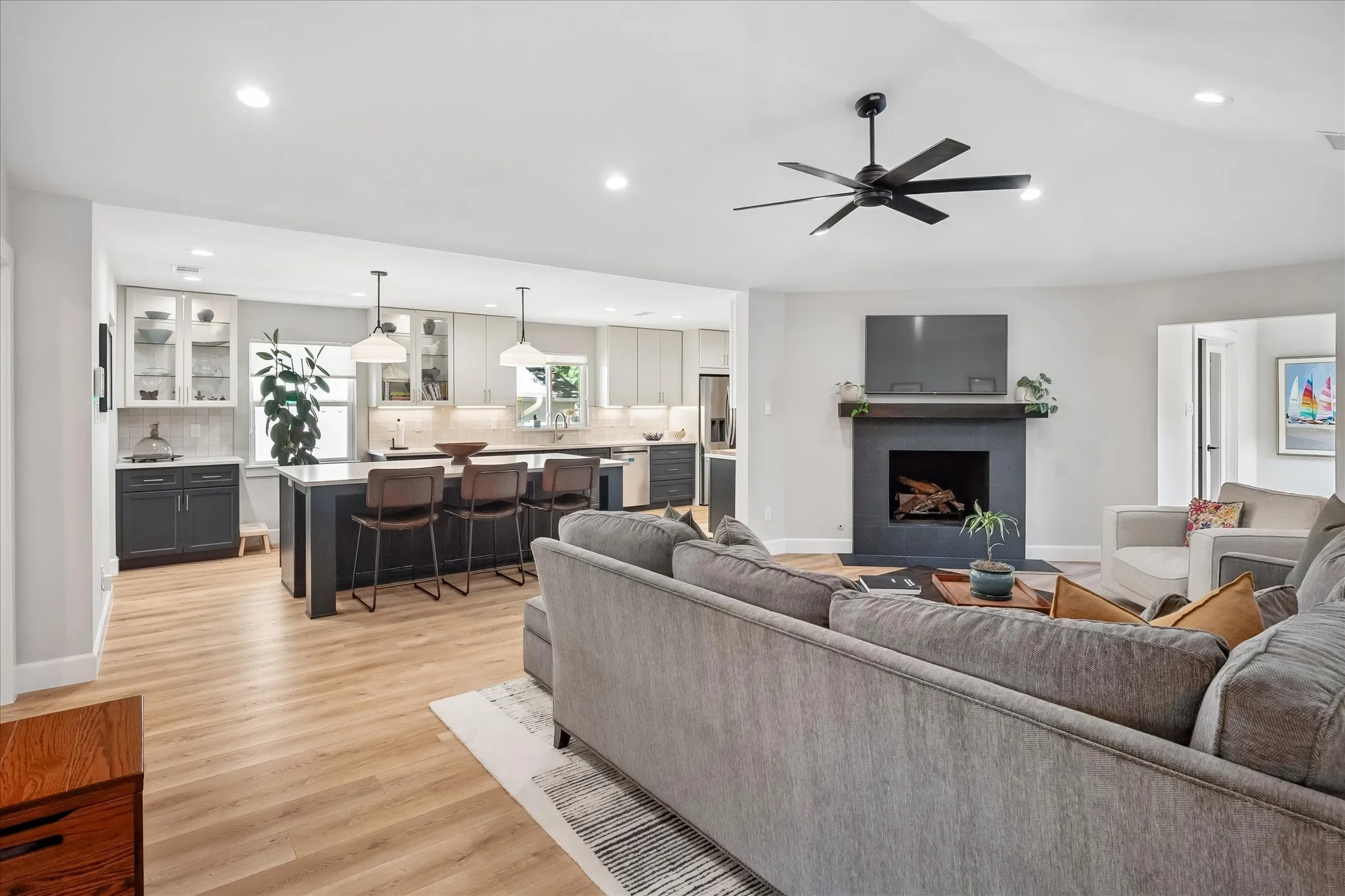 Open concept living area with wood-finish flooring, a fireplace with a dark mantel, a ceiling fan, a kitchen island with seating, and pendant lighting